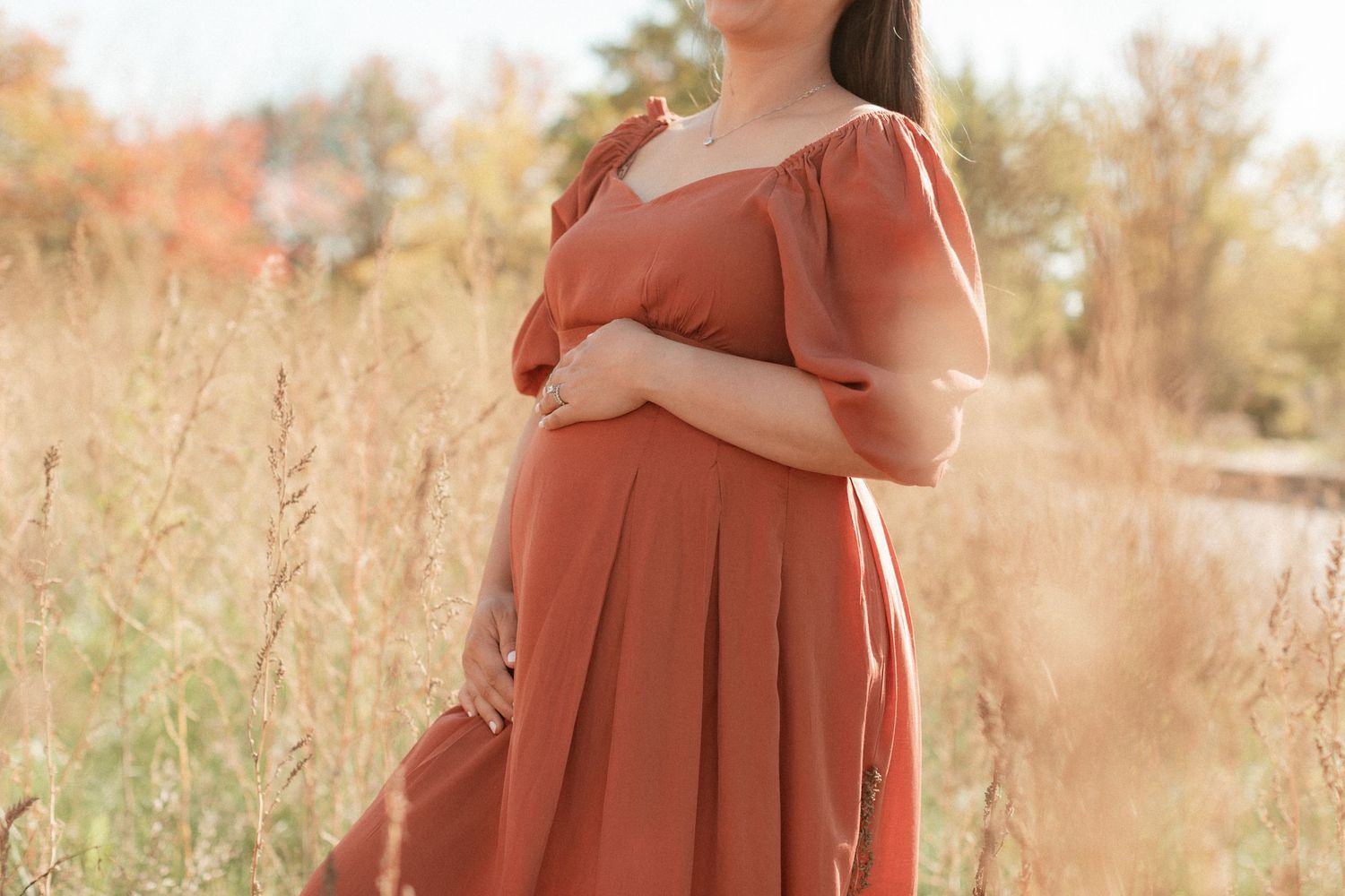 Side view of pregnant woman in terracotta dress standing among tall grass.