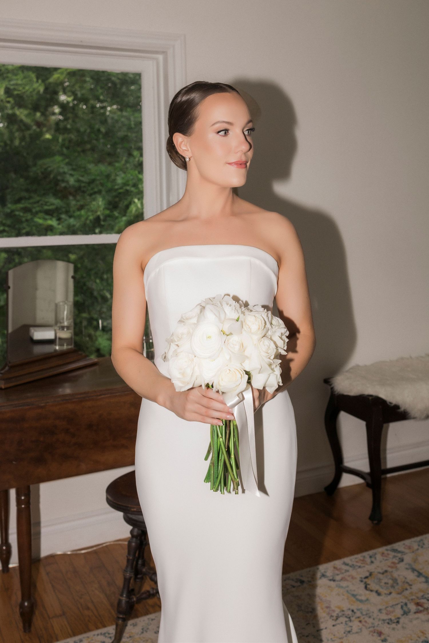 A classic strapless white wedding gown paired with a bouquet of white flowers next to a window.