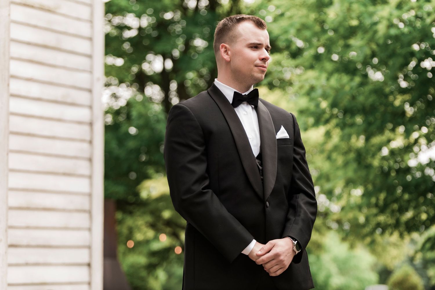 A groom stands in a black tuxedo and bow tie outdoors against a white building with greenery in the background.