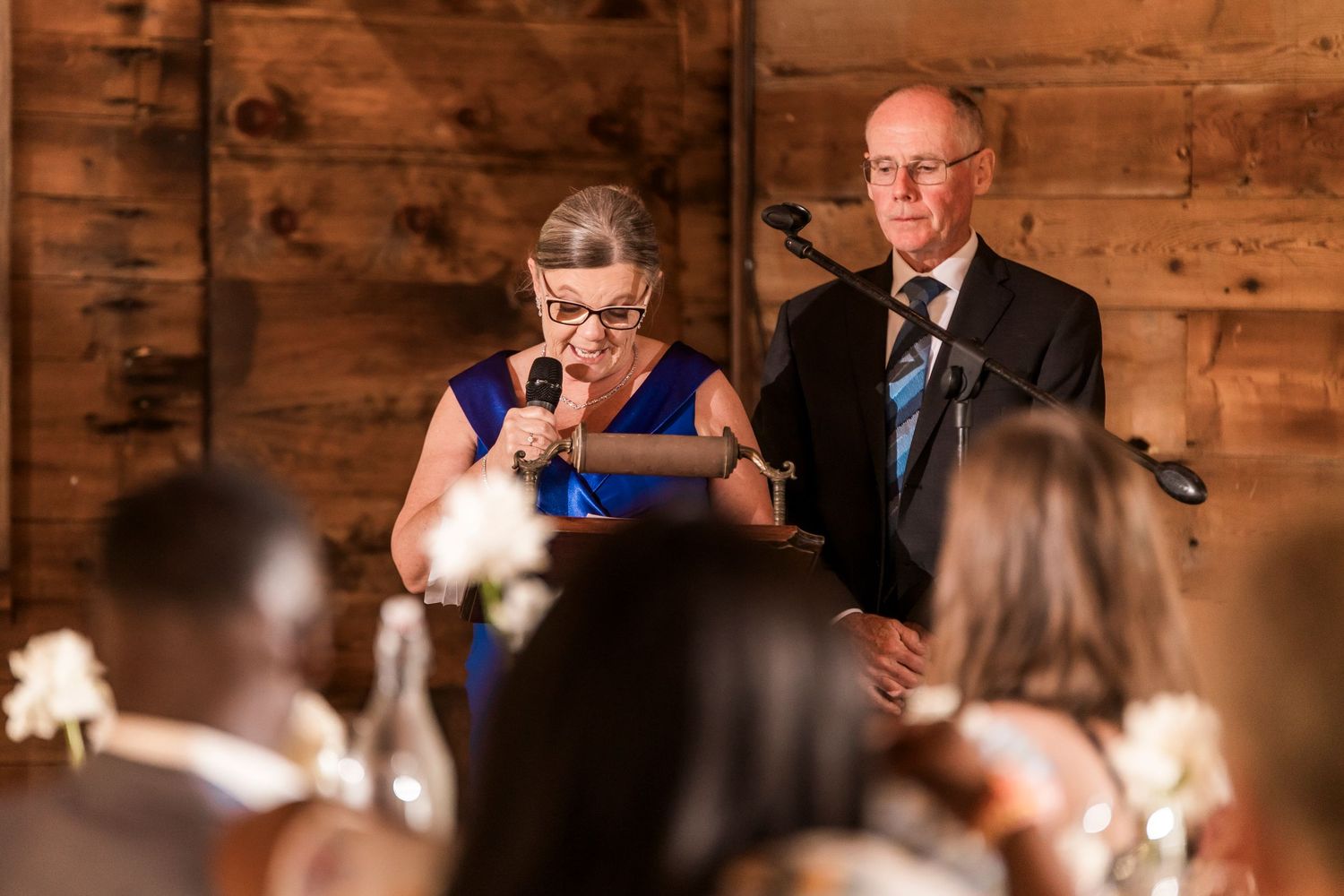 Speech is delivered to wedding guests in rustic wooden barn venue illuminated by candlelight.