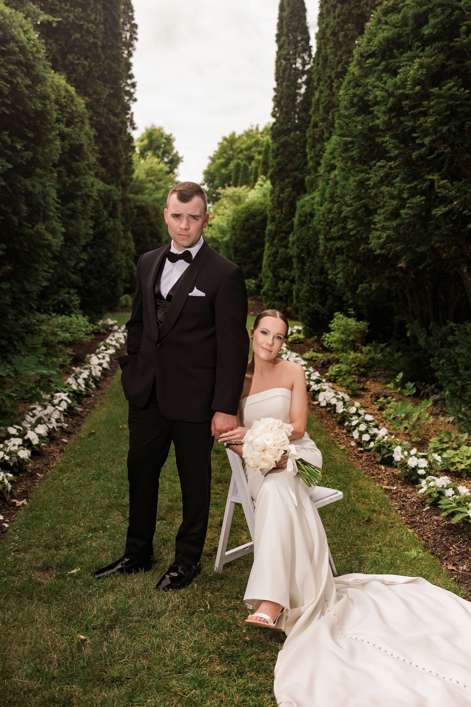 A romantic garden wedding scene with cypress trees and white floral arrangements lining the path.