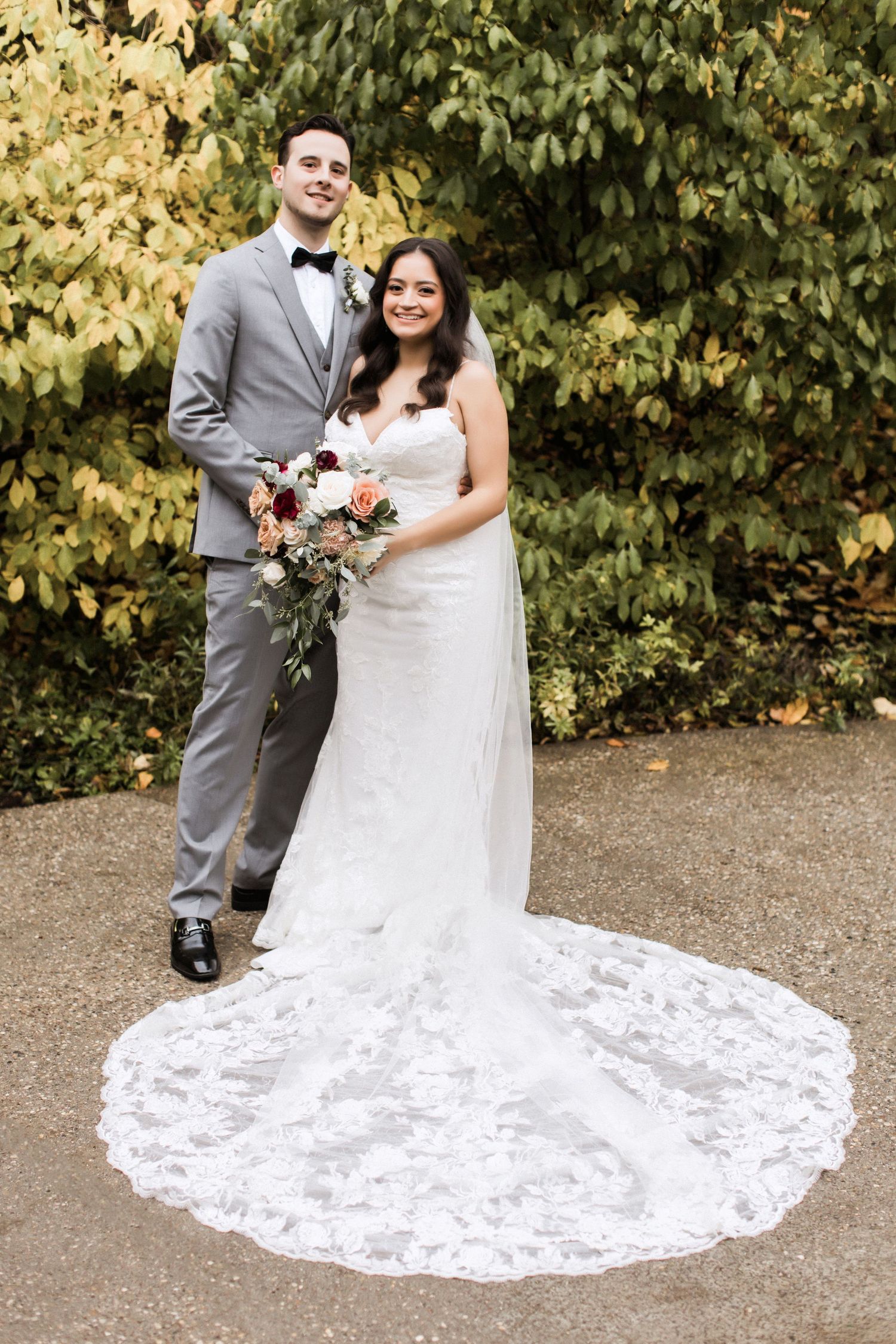 Couple poses in formal wedding attire with ornate lace train spread on pathway.