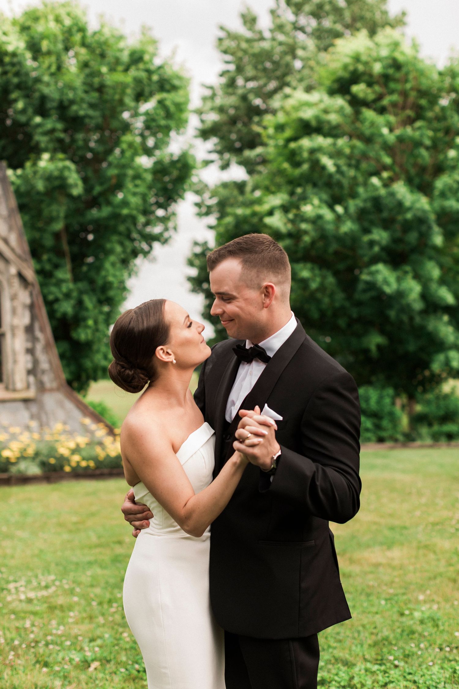 A romantic wedding couple shares joyful dance moves on a lush green lawn surrounded by trees.