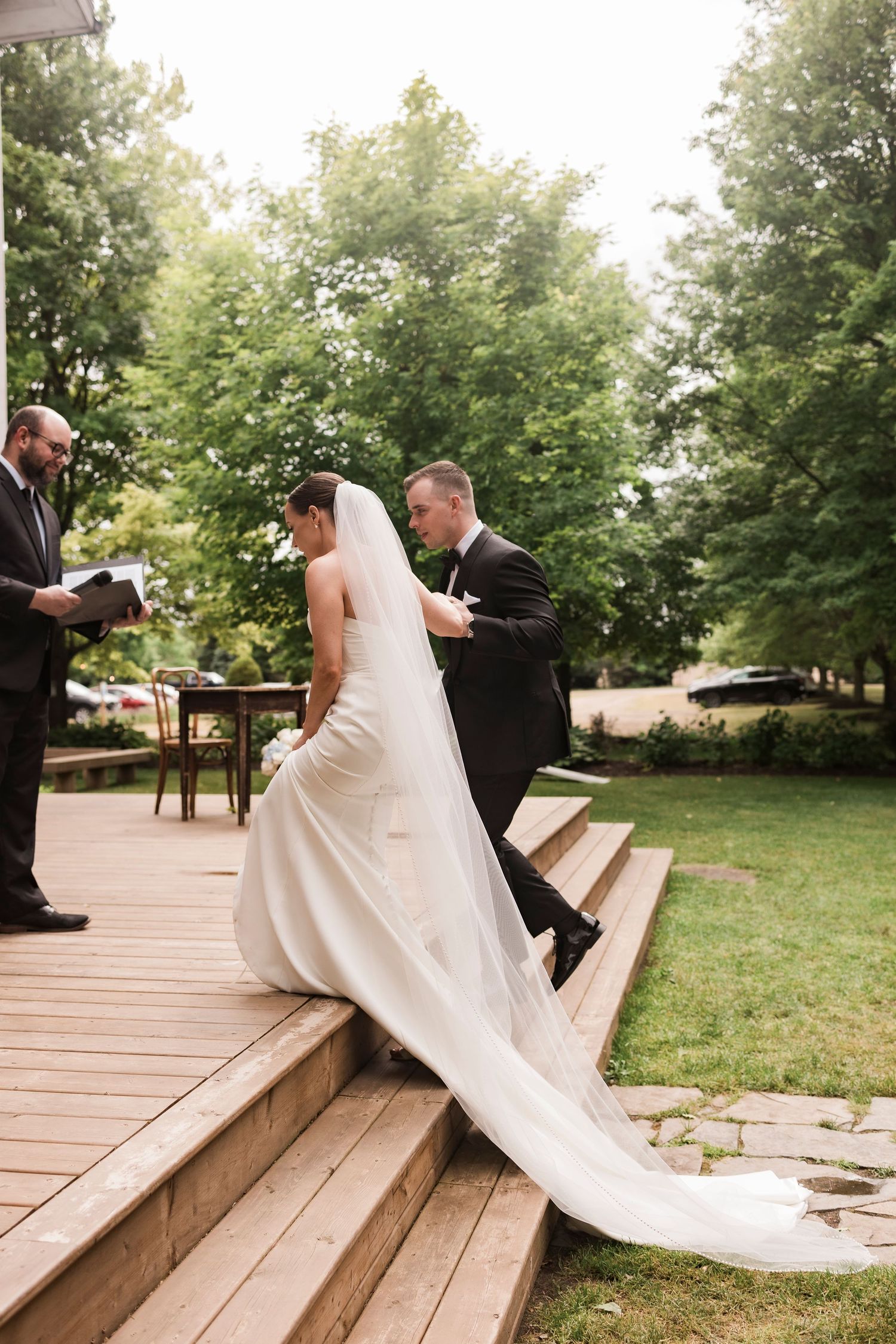 A couple walks up wooden deck stairs during their outdoor wedding ceremony as a trailing white dress flows behind.