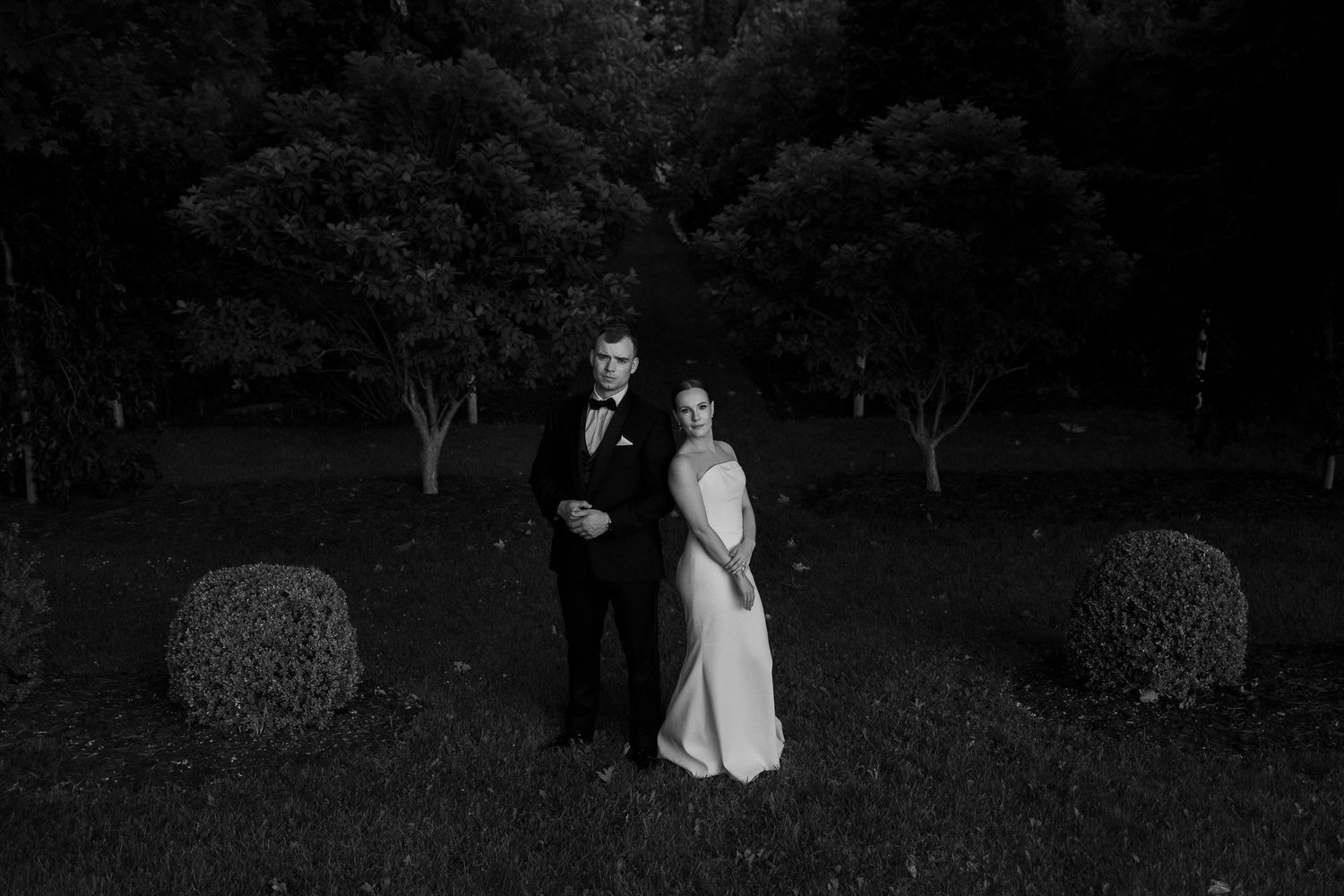 Elegant black and white photo of wedding couple standing in a garden with manicured topiary spheres at dusk.