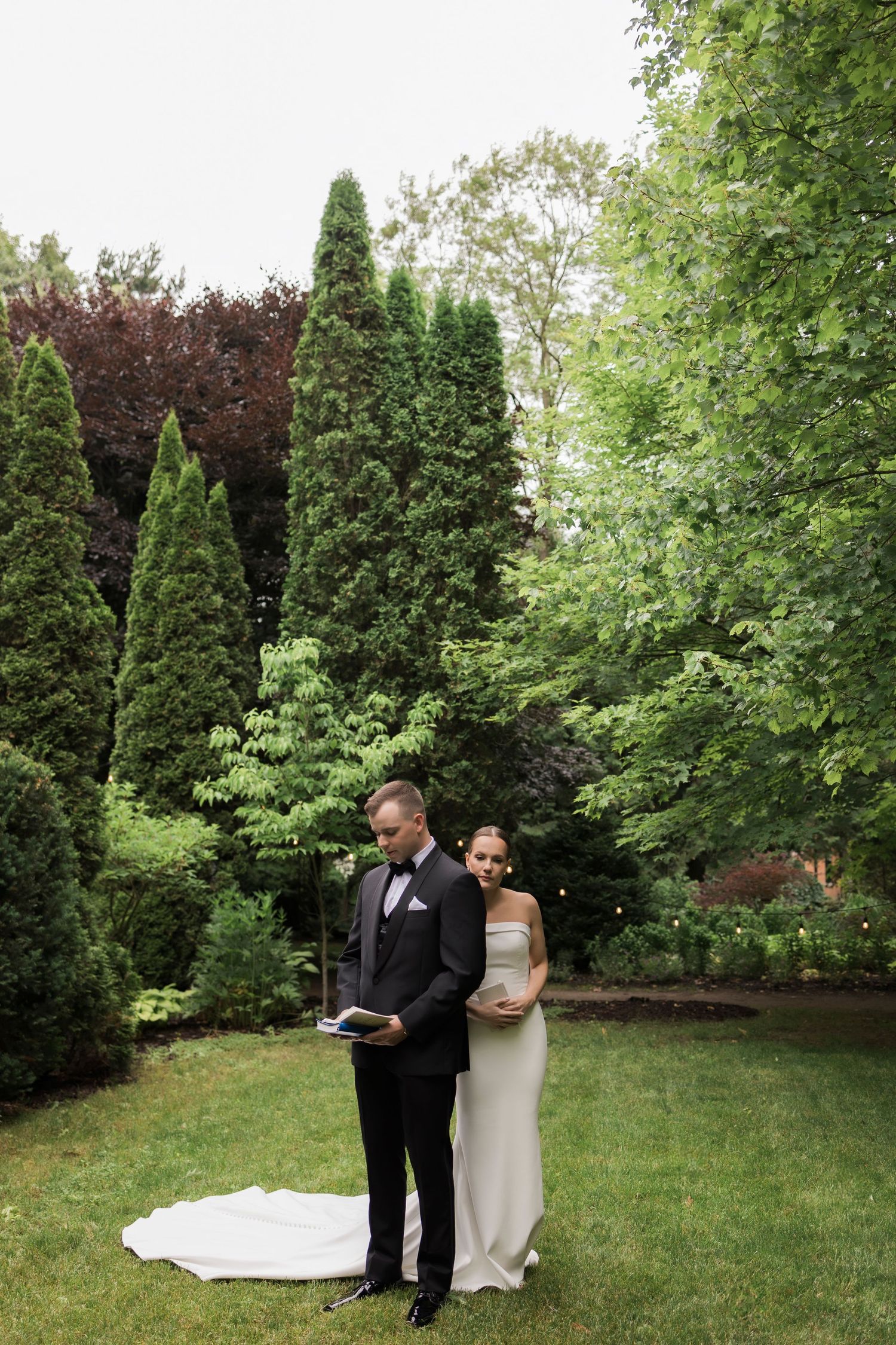 Wedding couple stands together in an elegant garden surrounded by tall evergreen trees.