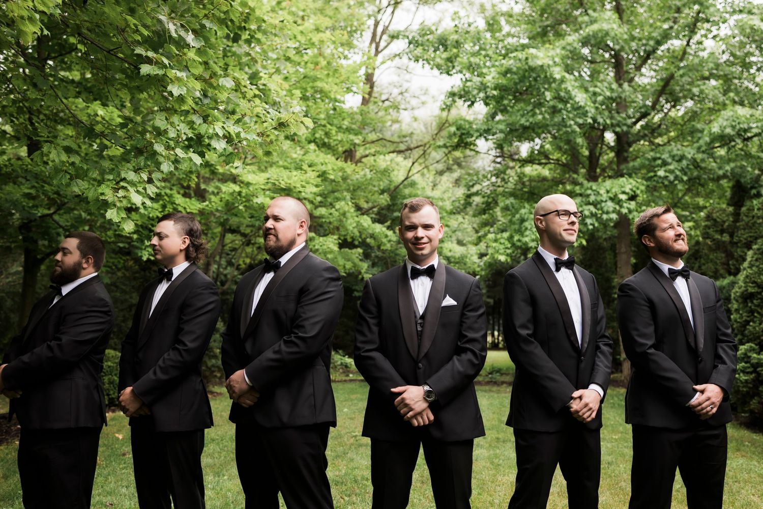 Black-tie wedding party lined up in formal tuxedos against a backdrop of green trees.