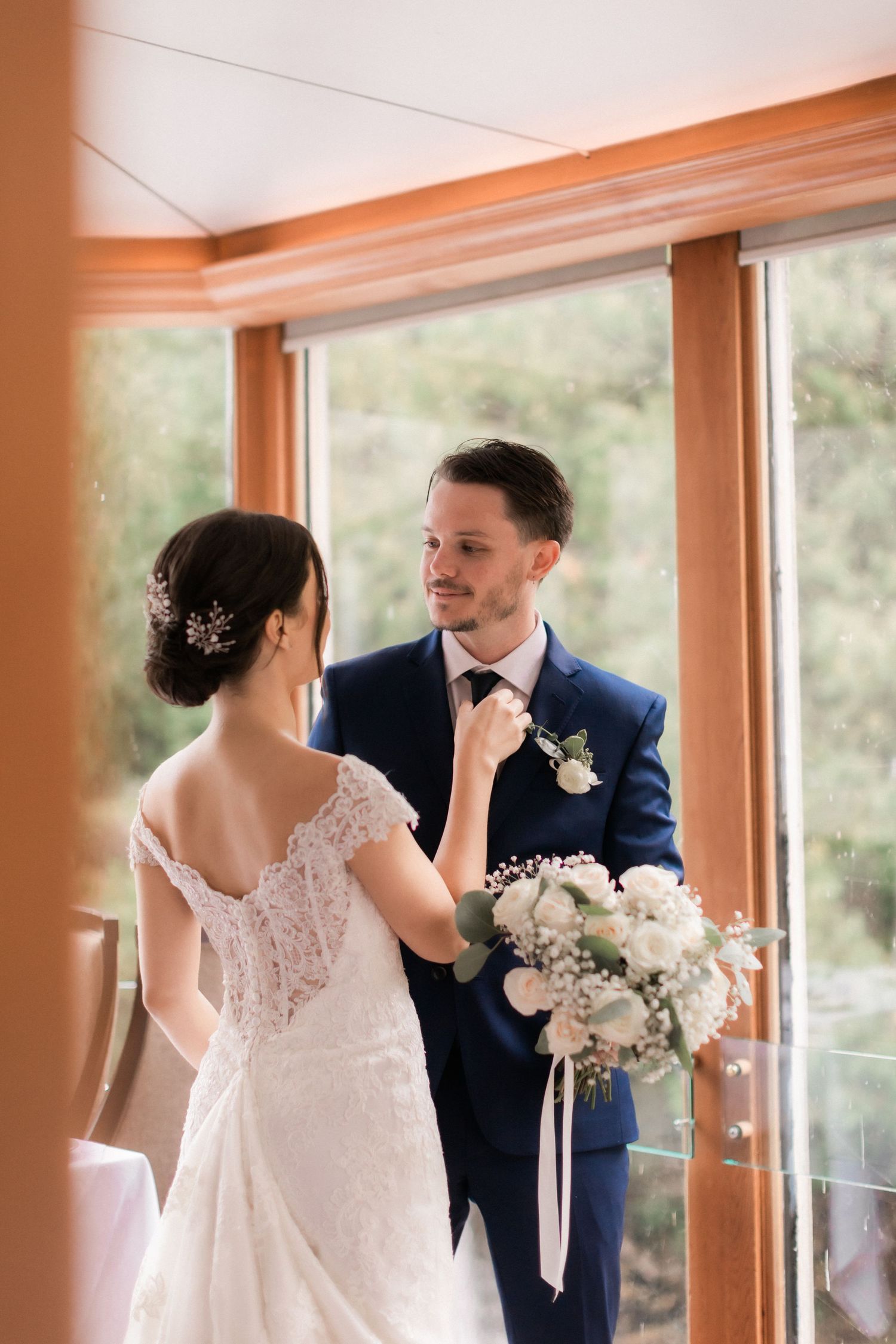 Bride adjusts groom's tie in intimate moment by window with natural light.
