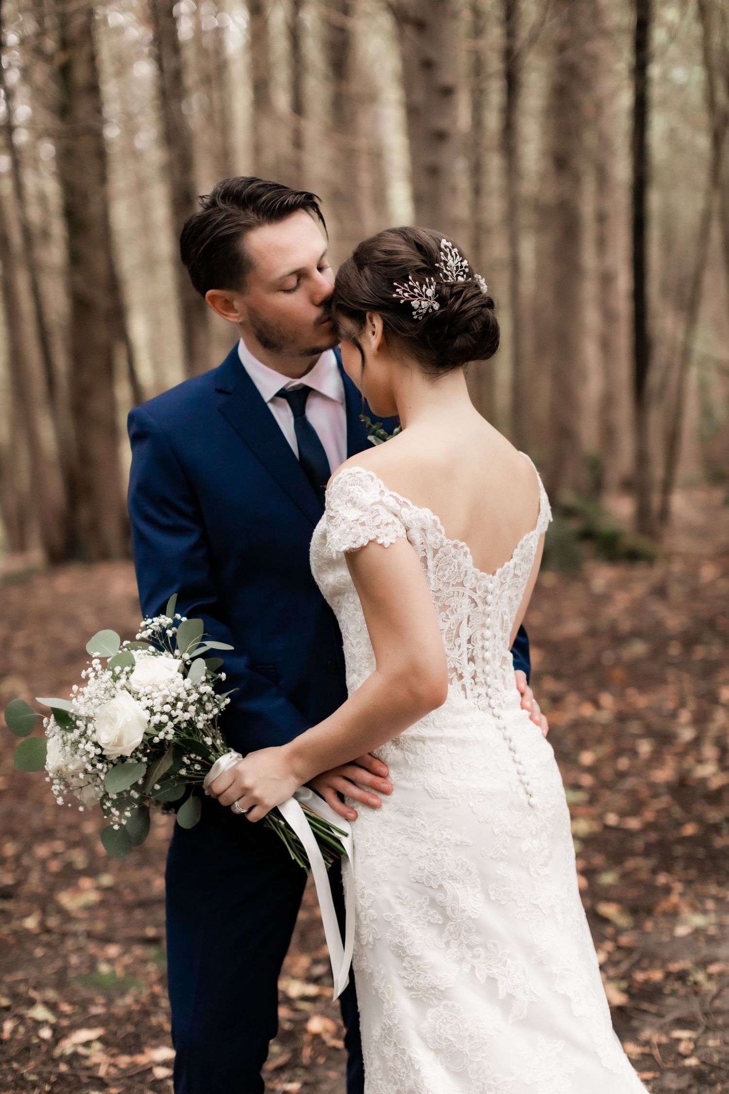 Intimate moment between newlyweds in a wooded setting, bride wearing lace dress with delicate back detail.