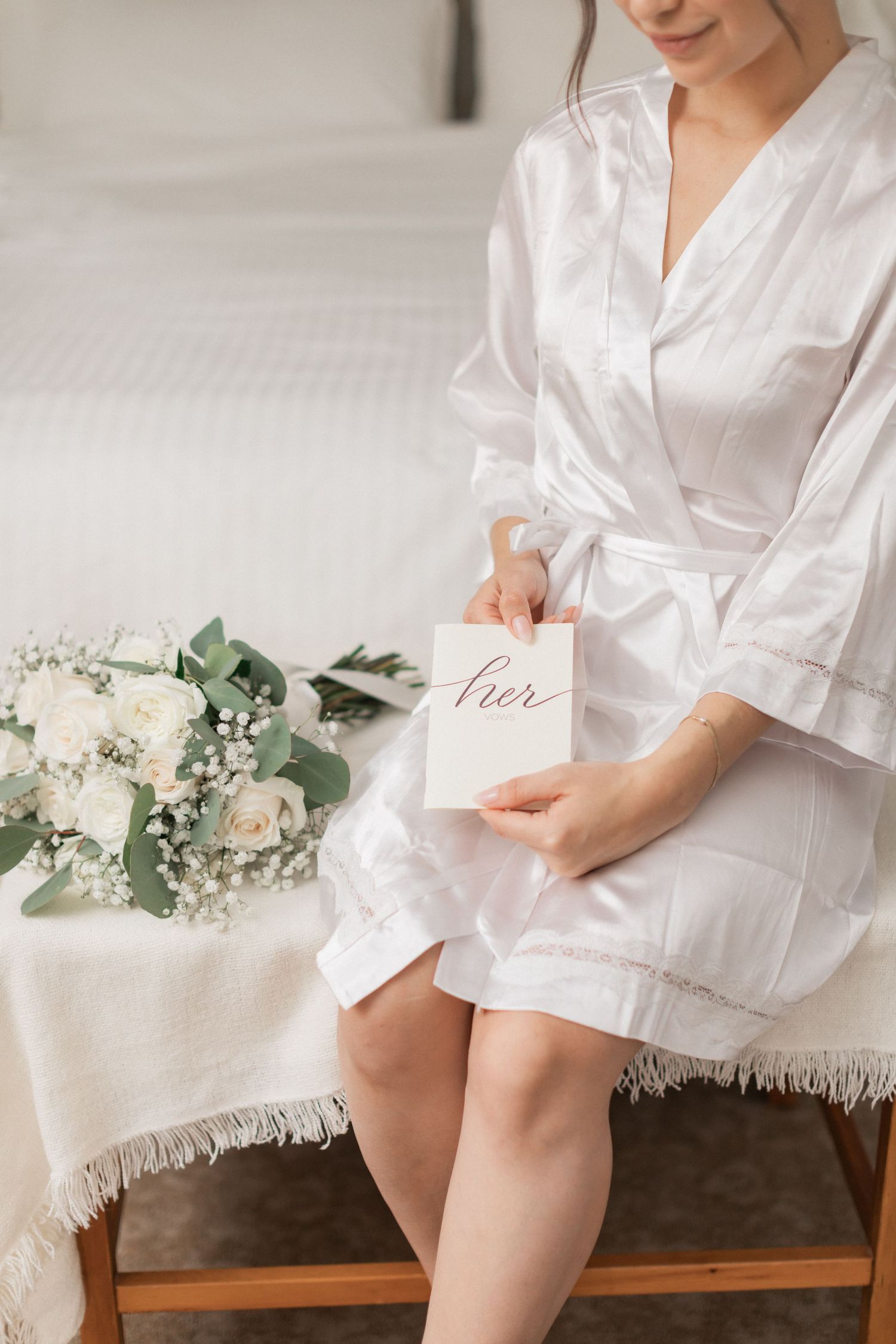 Bride in white silk robe holds wedding card with floral arrangement on bench.