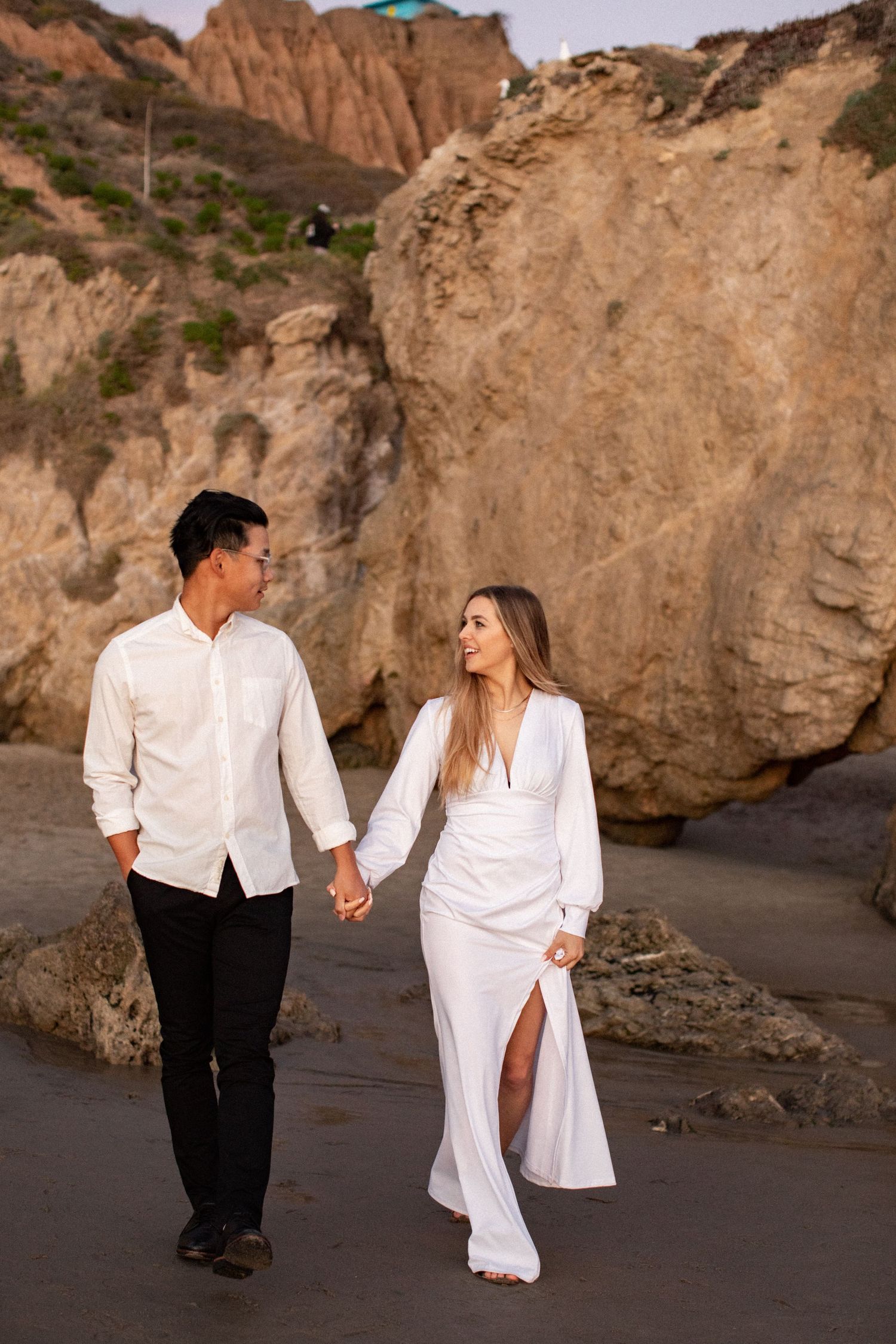 Couple strolling together on beach path with dramatic cliffs behind them.