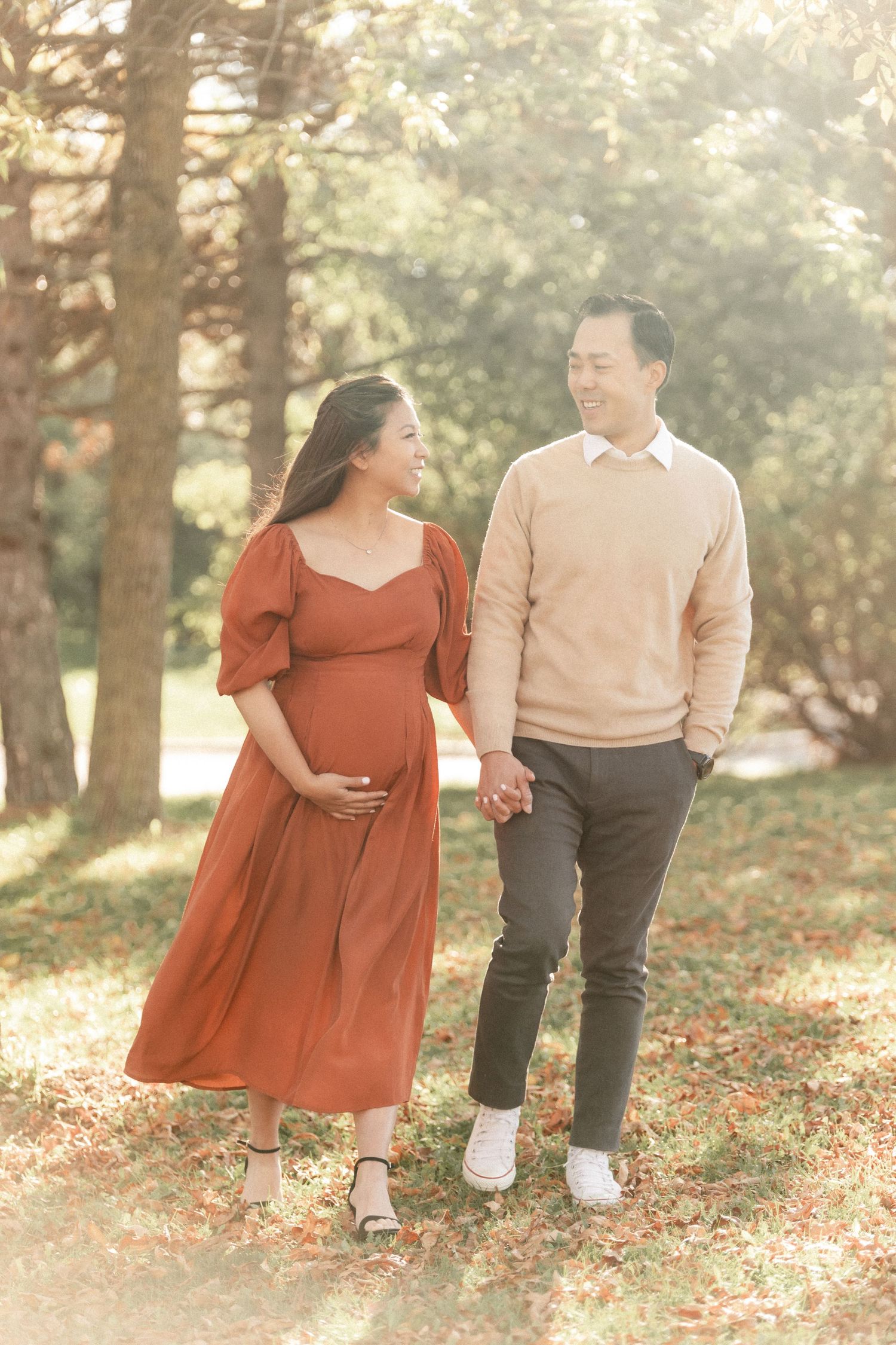 An expecting couple walks hand in hand through a wooded area in coordinated casual attire.