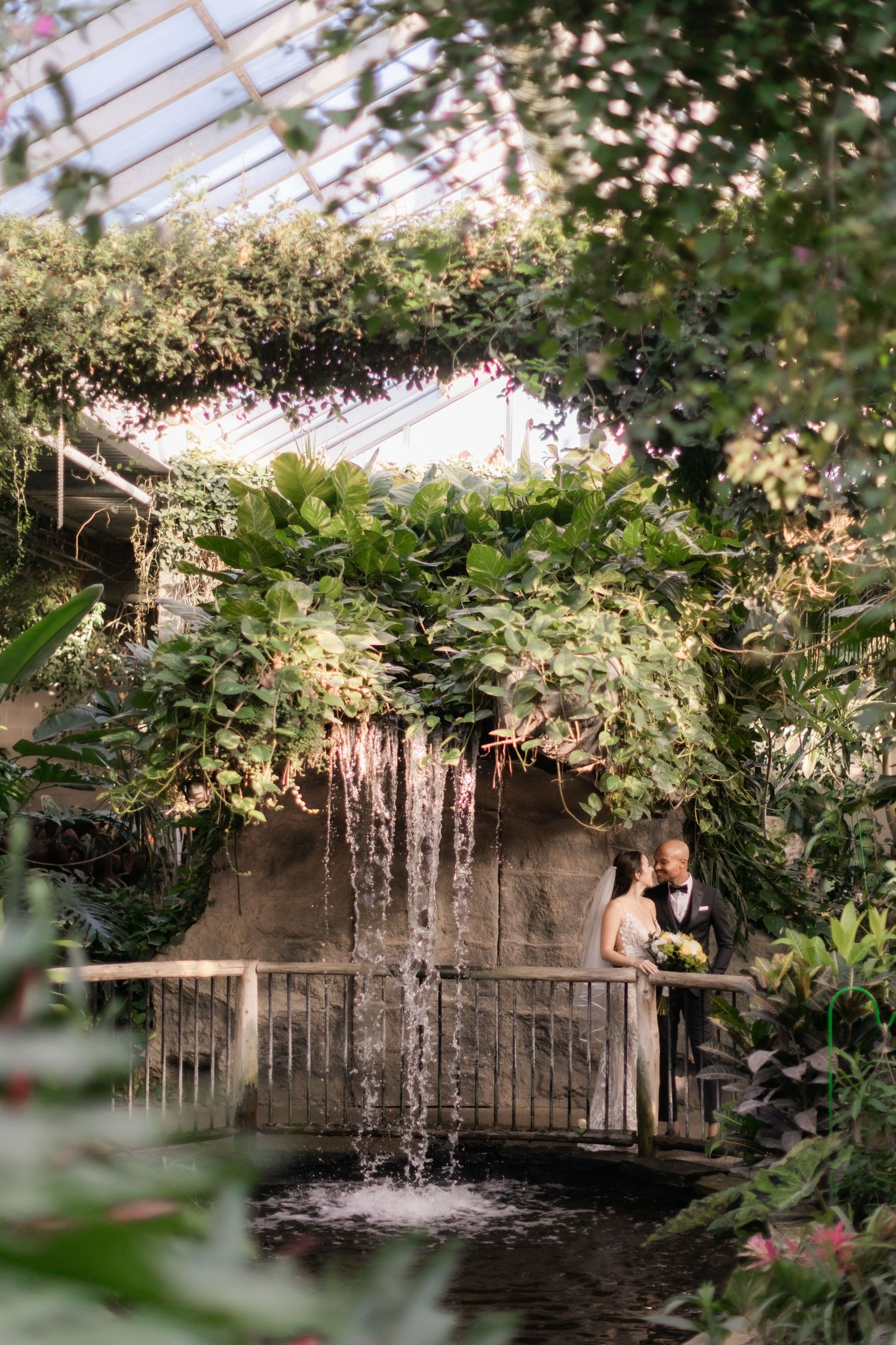 Romantic garden wedding photo with couple framed by waterfall and hanging plants.