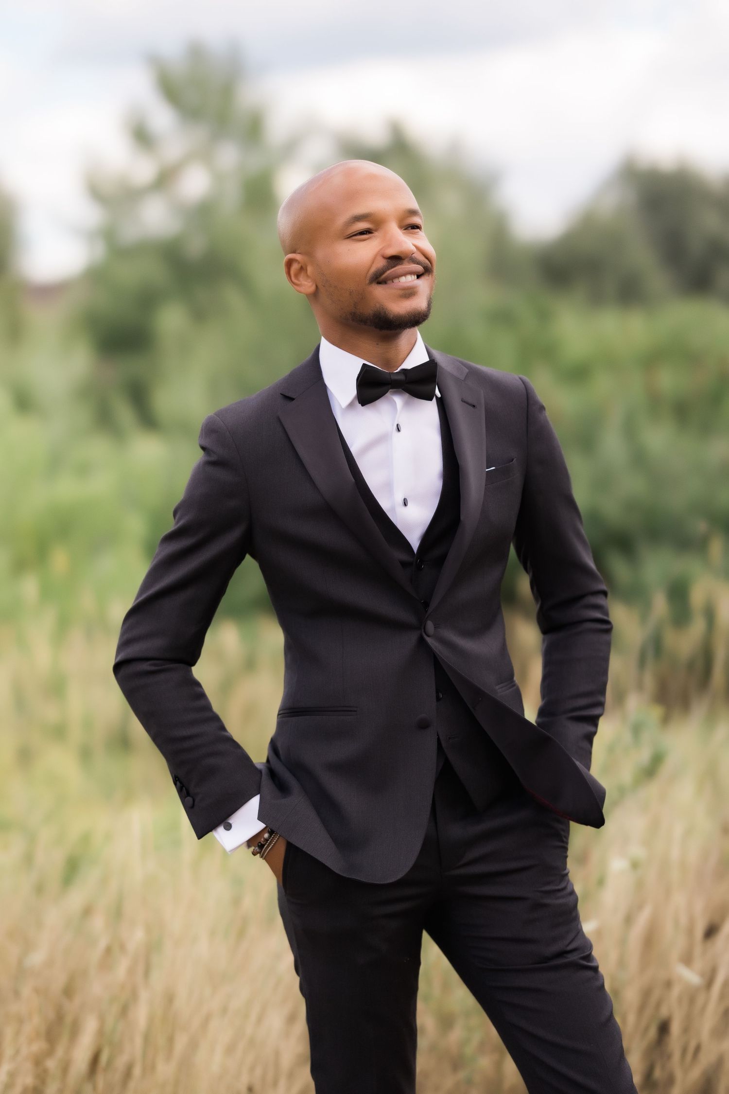 Groom in tailored black tuxedo poses confidently in outdoor field setting.