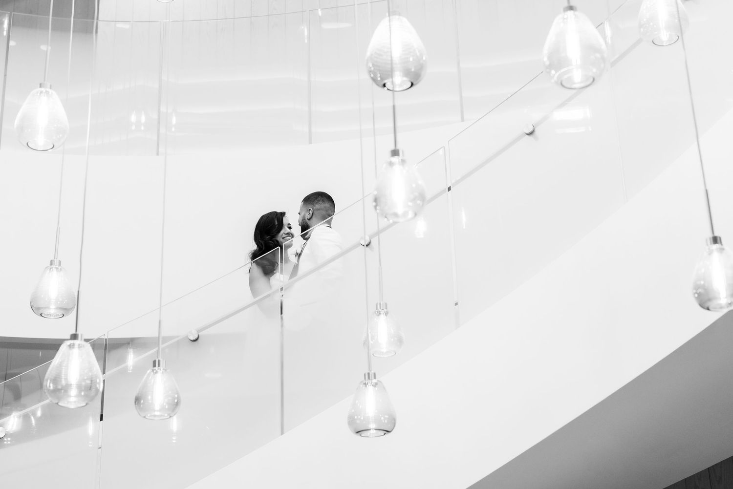 Black and white wedding photo showing couple on curving staircase beneath pendant lights.