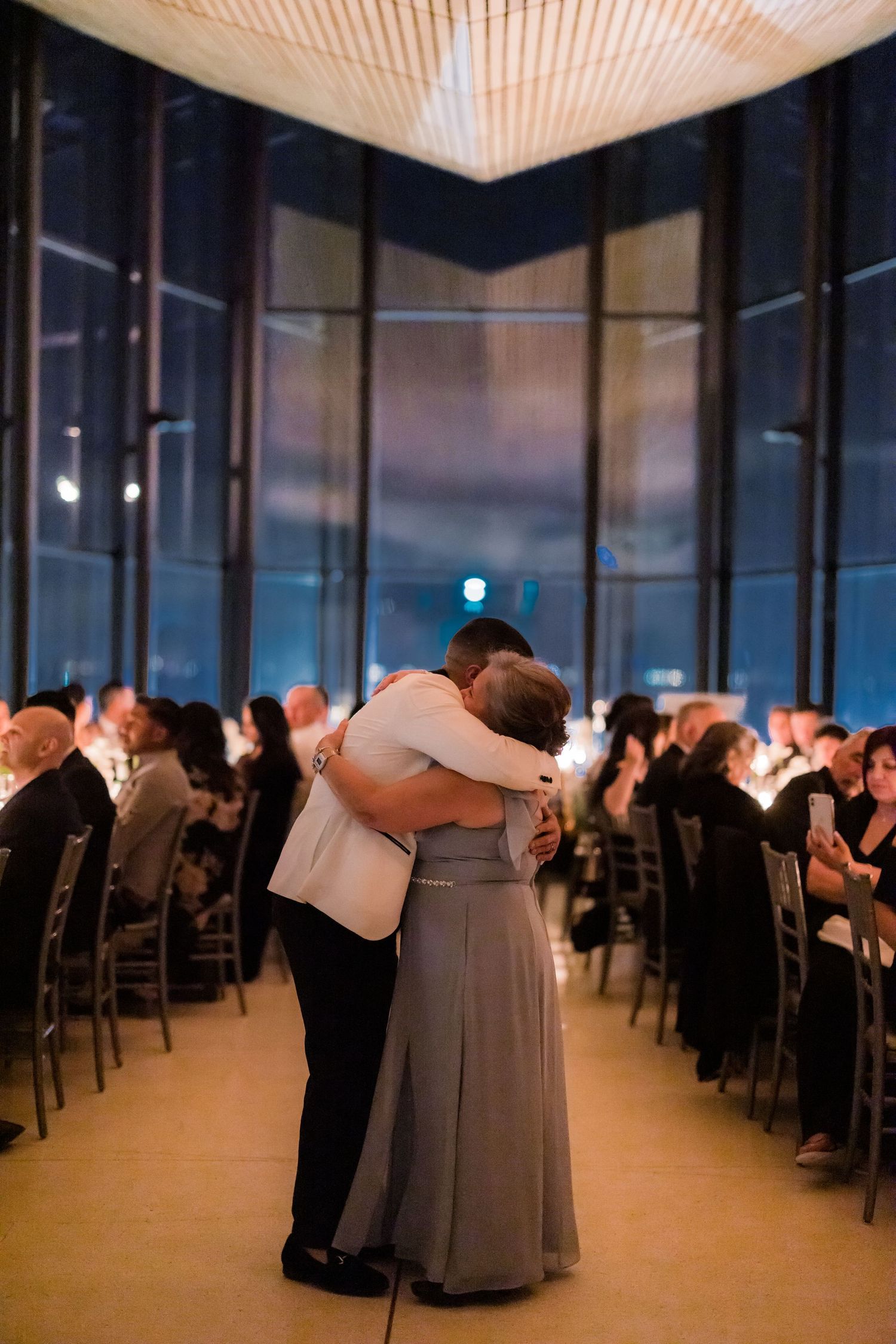 A romantic slow dance in a modern venue with floor-to-ceiling windows and candlelit reception tables.