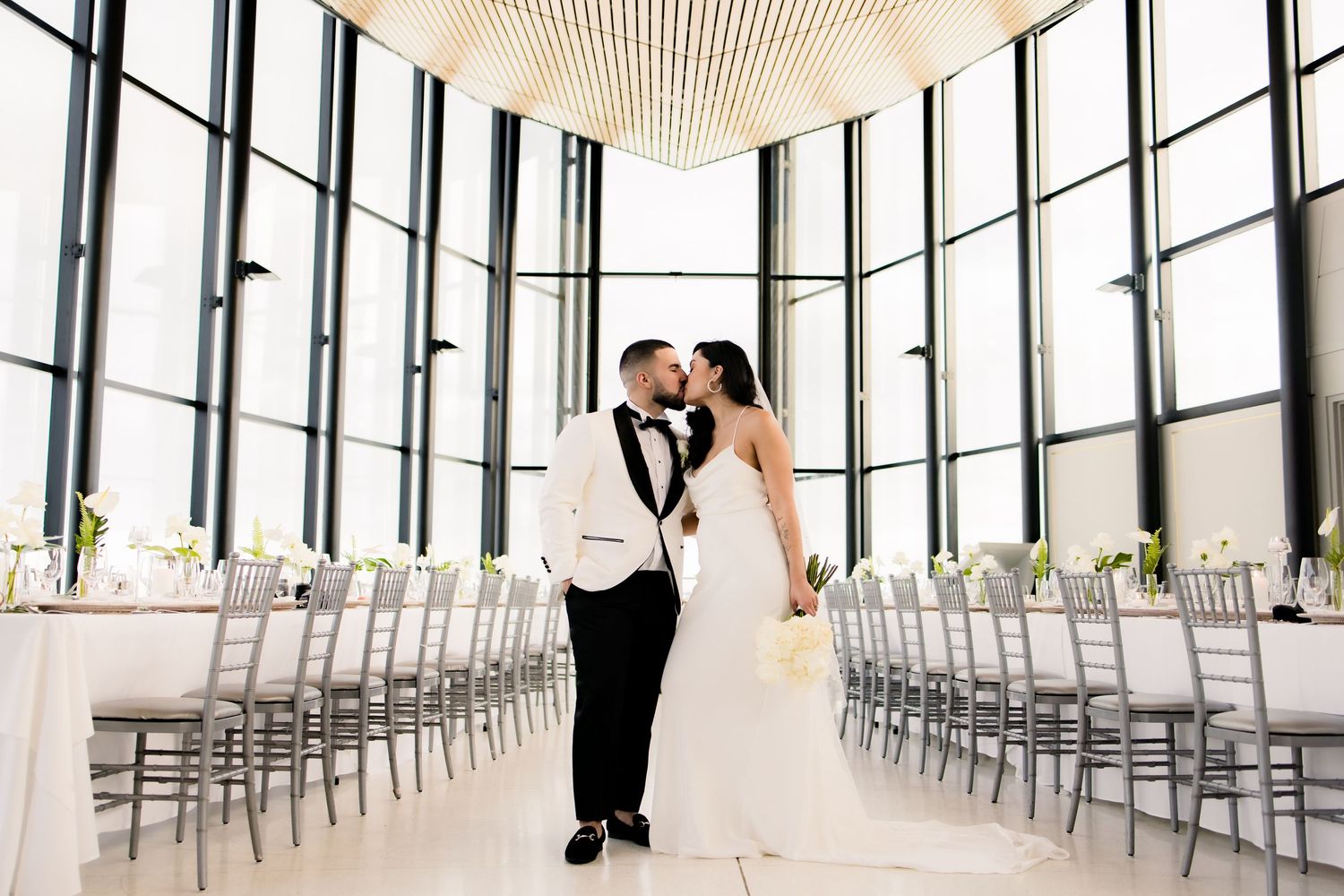 Wedding couple posing in modern venue with floor-to-ceiling windows and silver chiavari chairs.