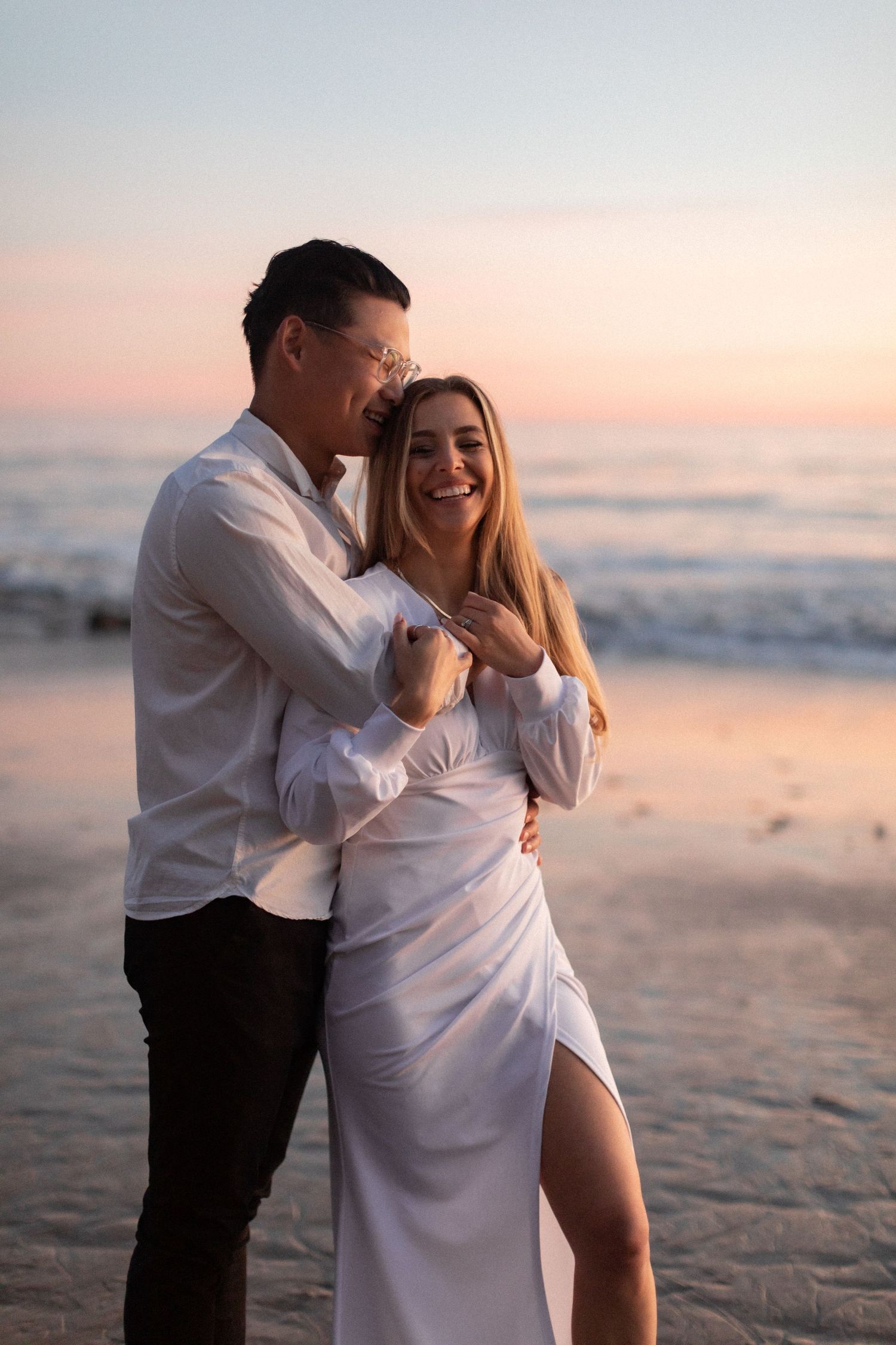 Couple embracing in white outfits during golden hour on a beach.