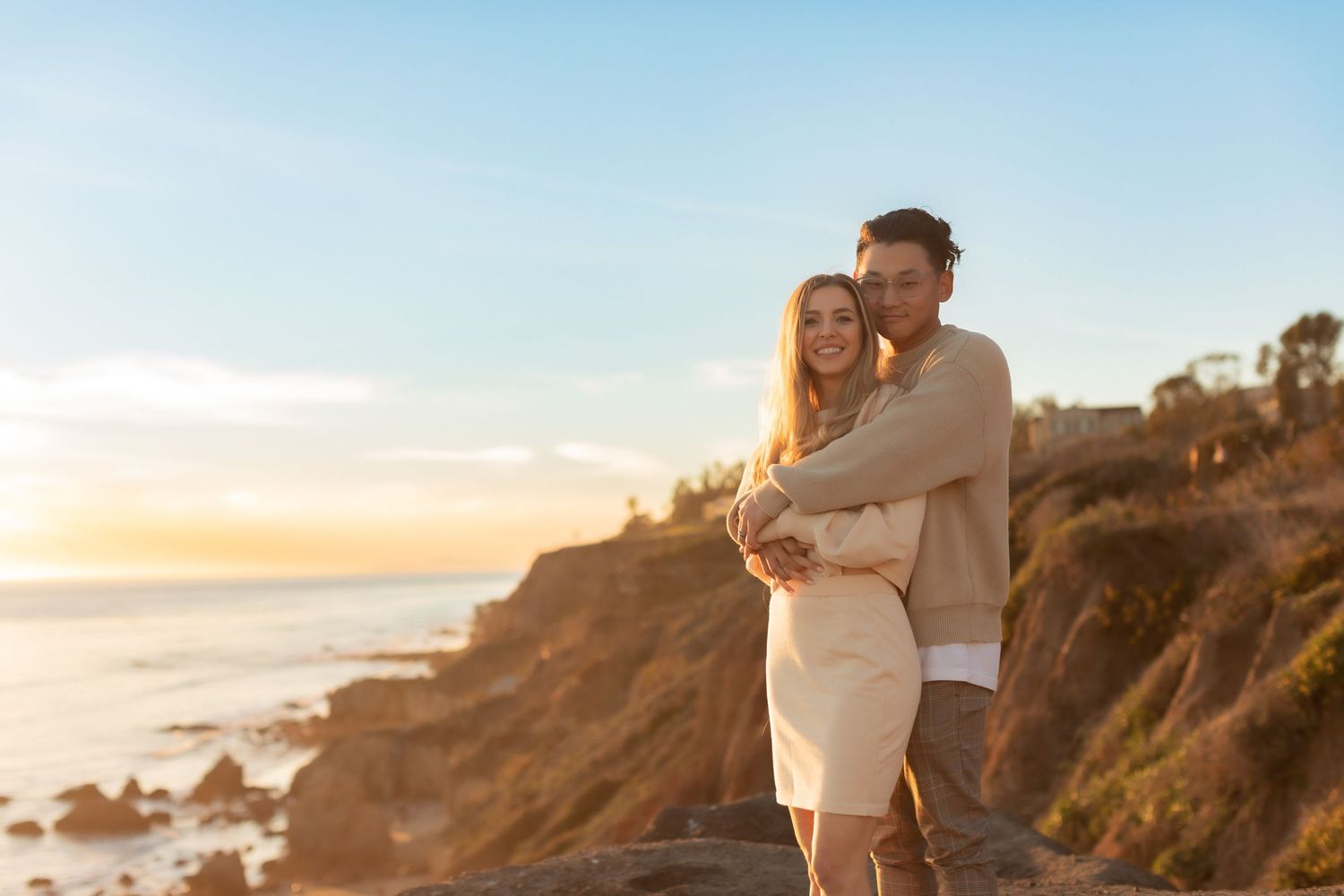 Couple in light-colored clothing embracing on rocky coastline during sunset.