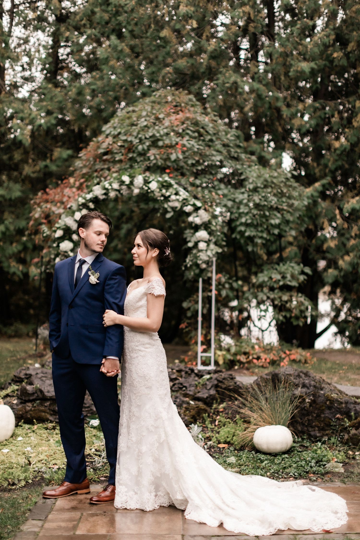 Elegant wedding couple standing under floral arch on brick pathway with white pumpkin decorations.