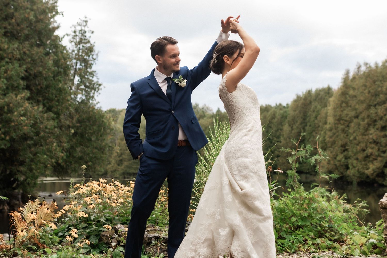 Couple dancing together outdoors near lake with golden flowers and greenery in background.