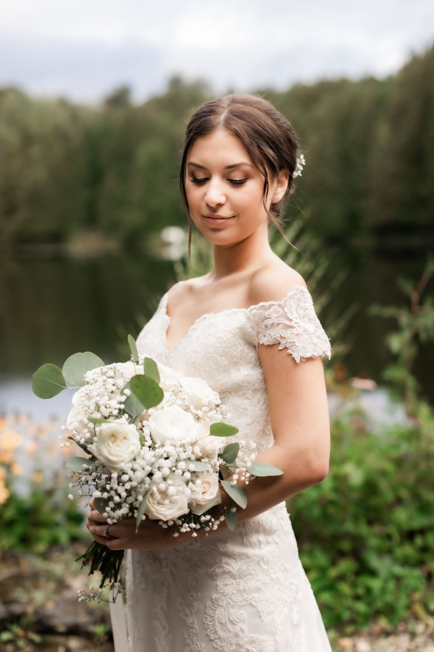 A bride in lace holds a delicate white and green bouquet by a lakeside.