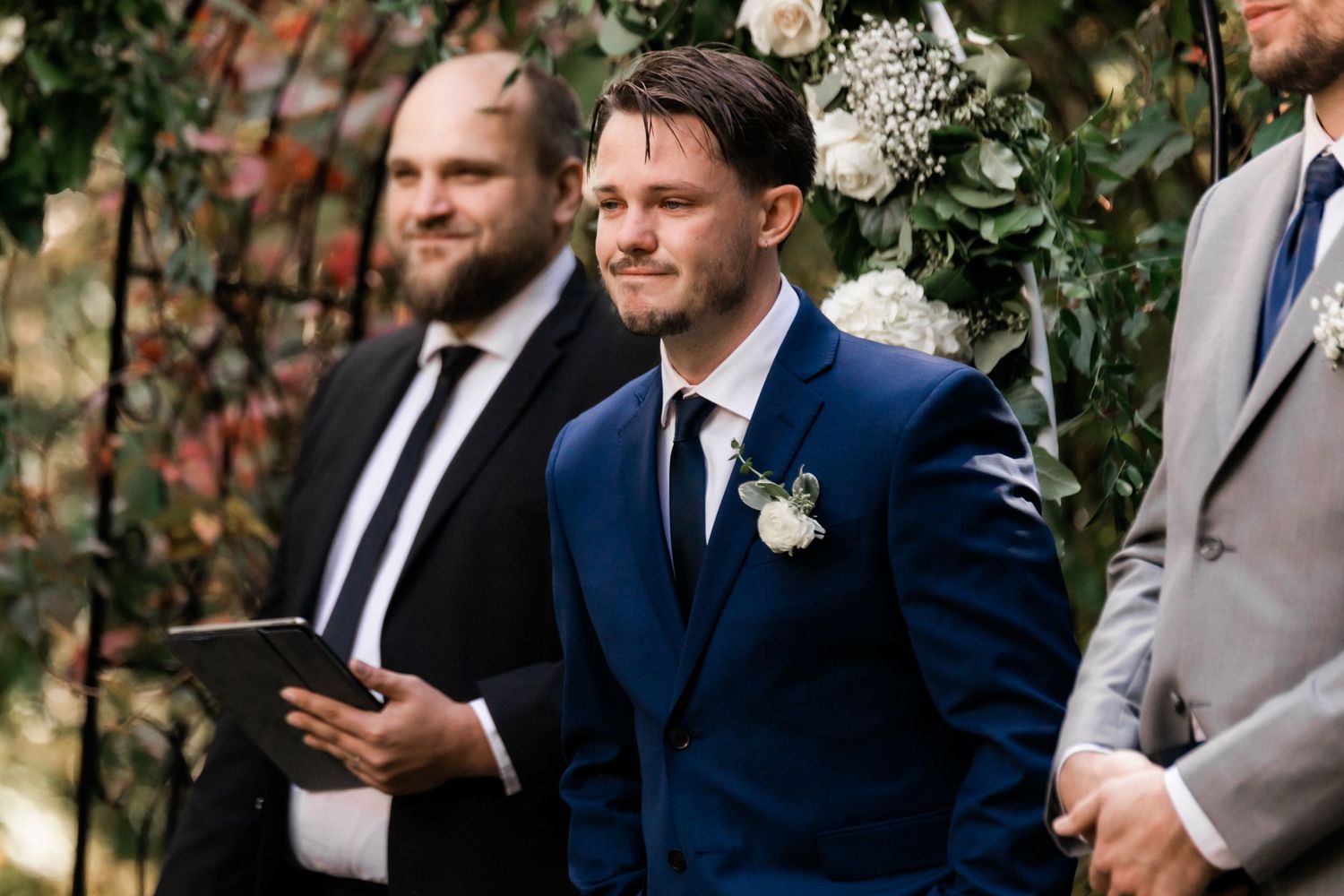 A groom in a navy suit stands with groomsmen during an outdoor garden ceremony.