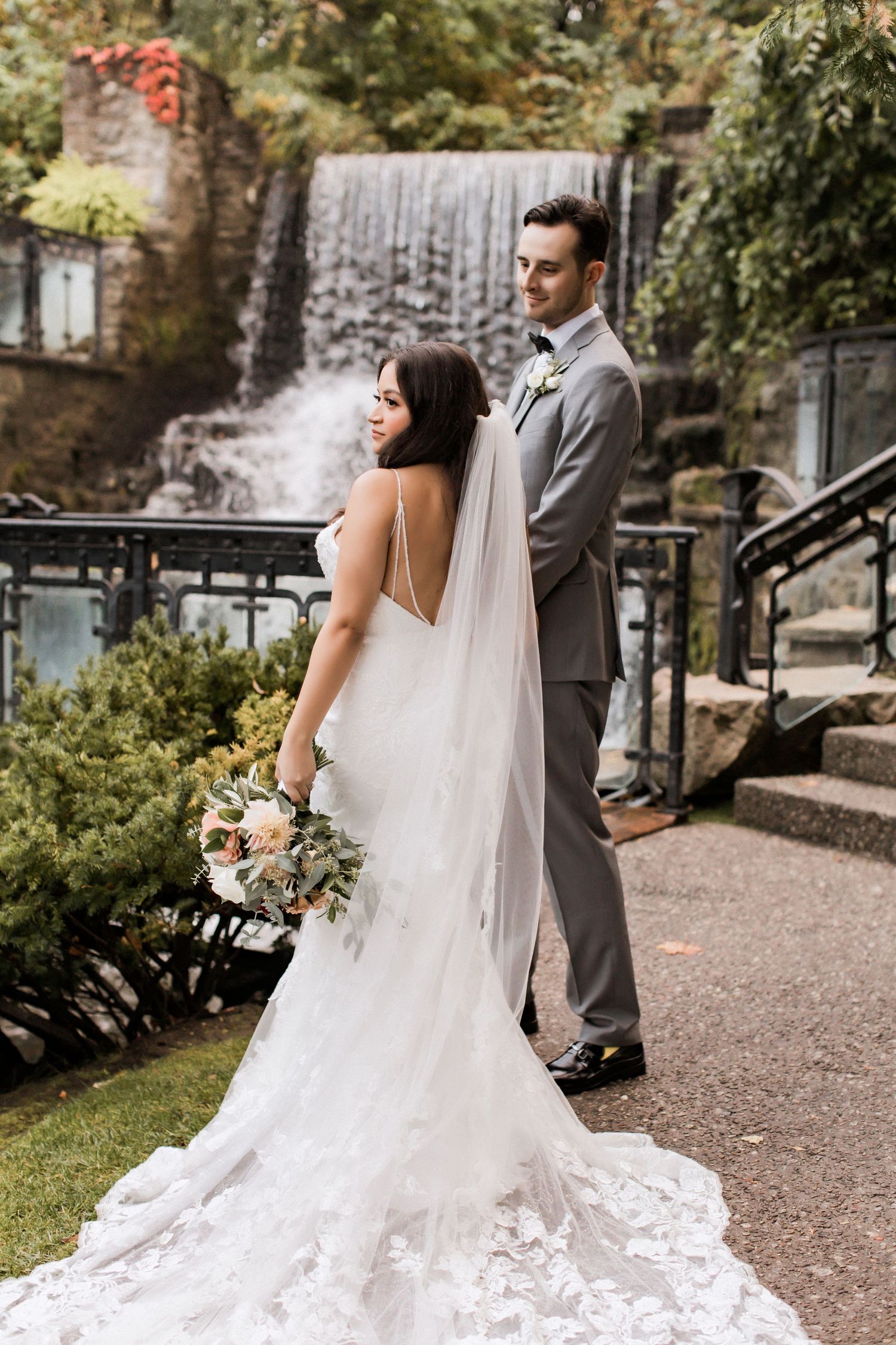 Bride in stunning lace gown stands with groom near garden waterfall feature.