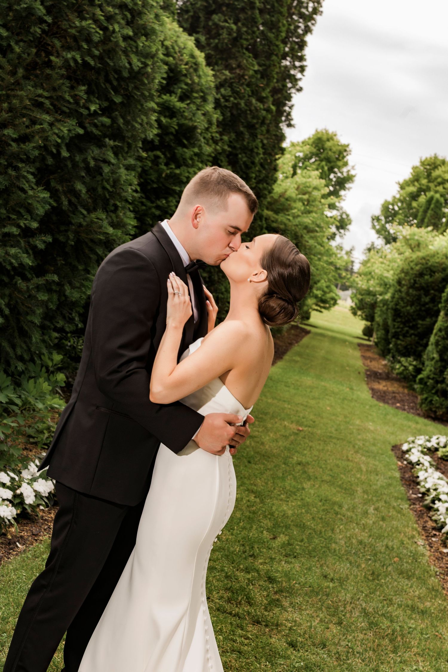 Romantic garden wedding photo of couple embracing on manicured lawn path with tall hedges.