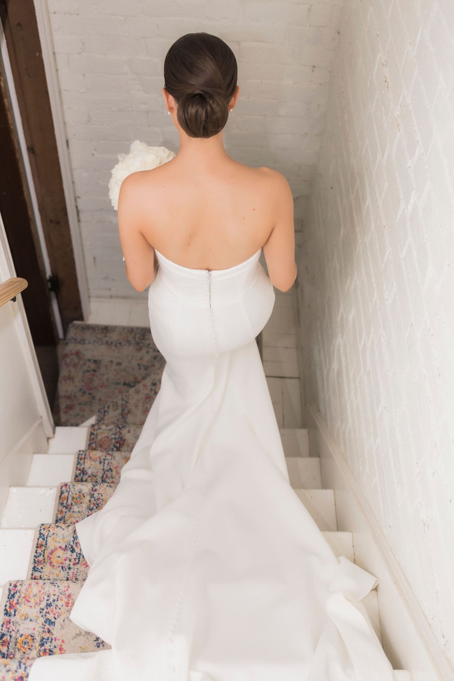 A bride in a form-fitting white wedding gown stands on carpeted stairs with her back to the camera.