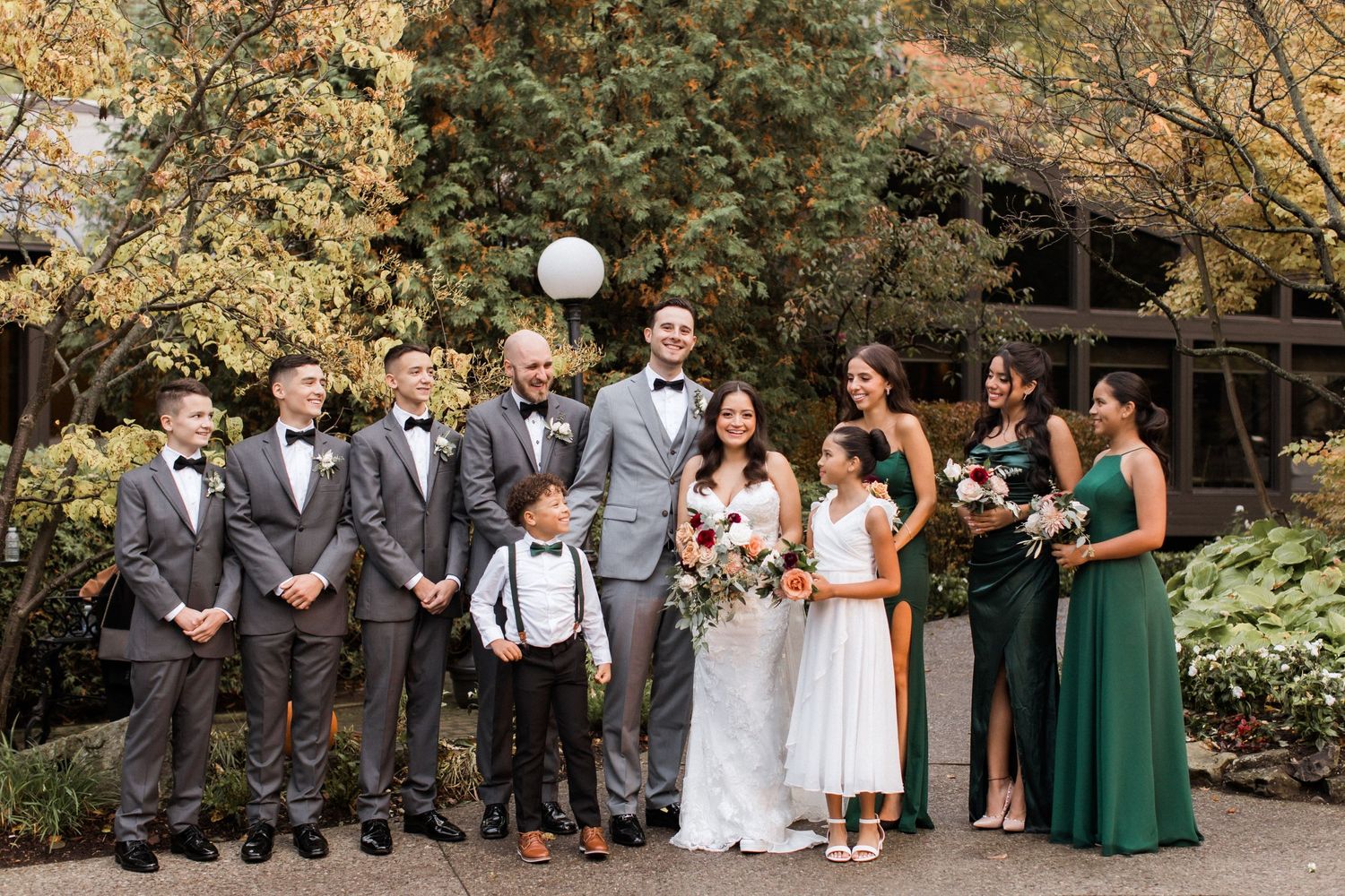Wedding party in grey suits and green dresses poses together on garden path with autumn foliage in background.