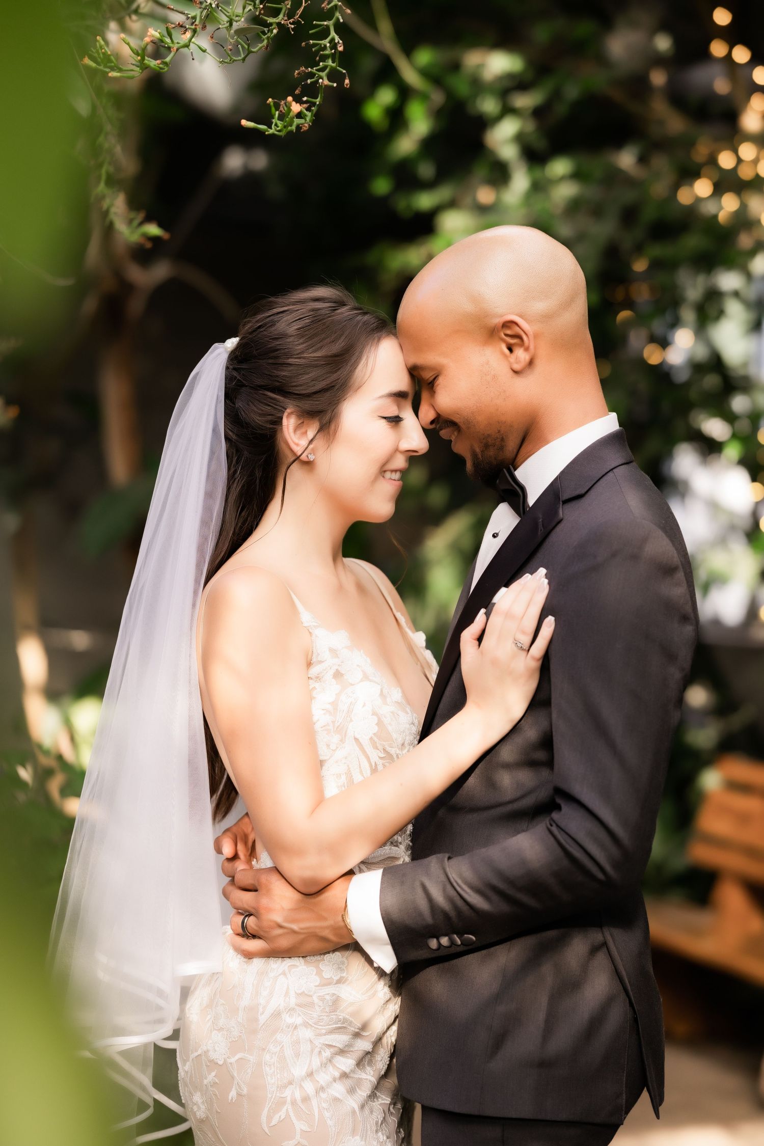 Intimate forehead touch between newlyweds surrounded by tropical foliage and string lights.