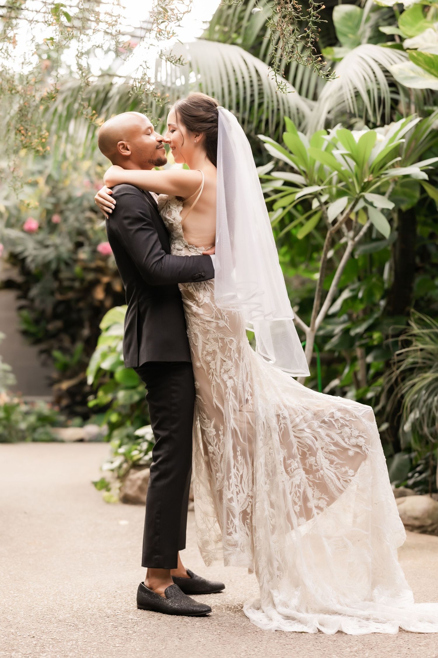 Full-length wedding portrait showing couple embracing in front of tropical greenhouse plants.
