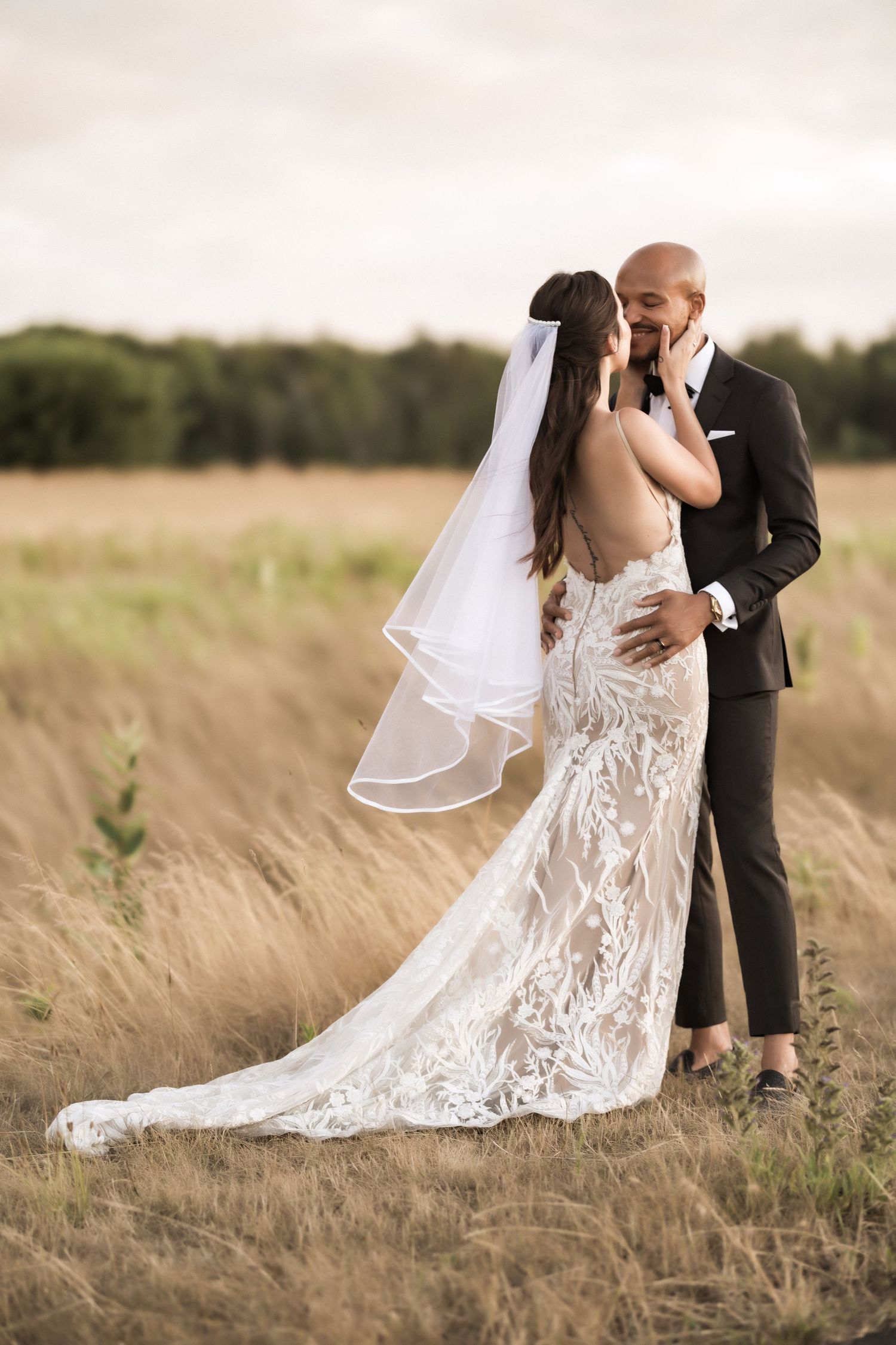 Bride in lace dress and groom share intimate moment in golden wheat field at sunset.