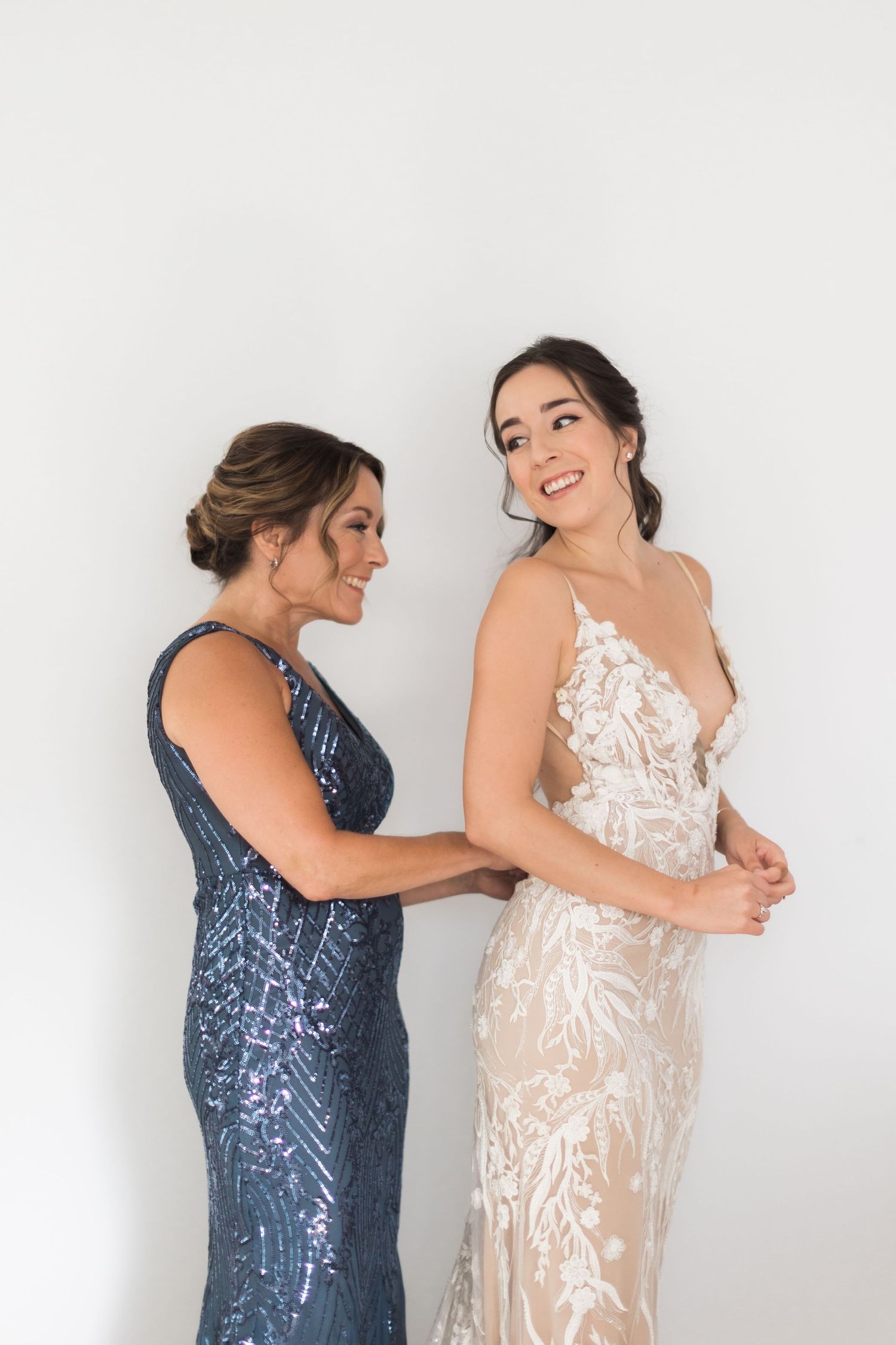 Two women share a joyful moment in elegant gowns, one wearing navy sequins and one in white lace.