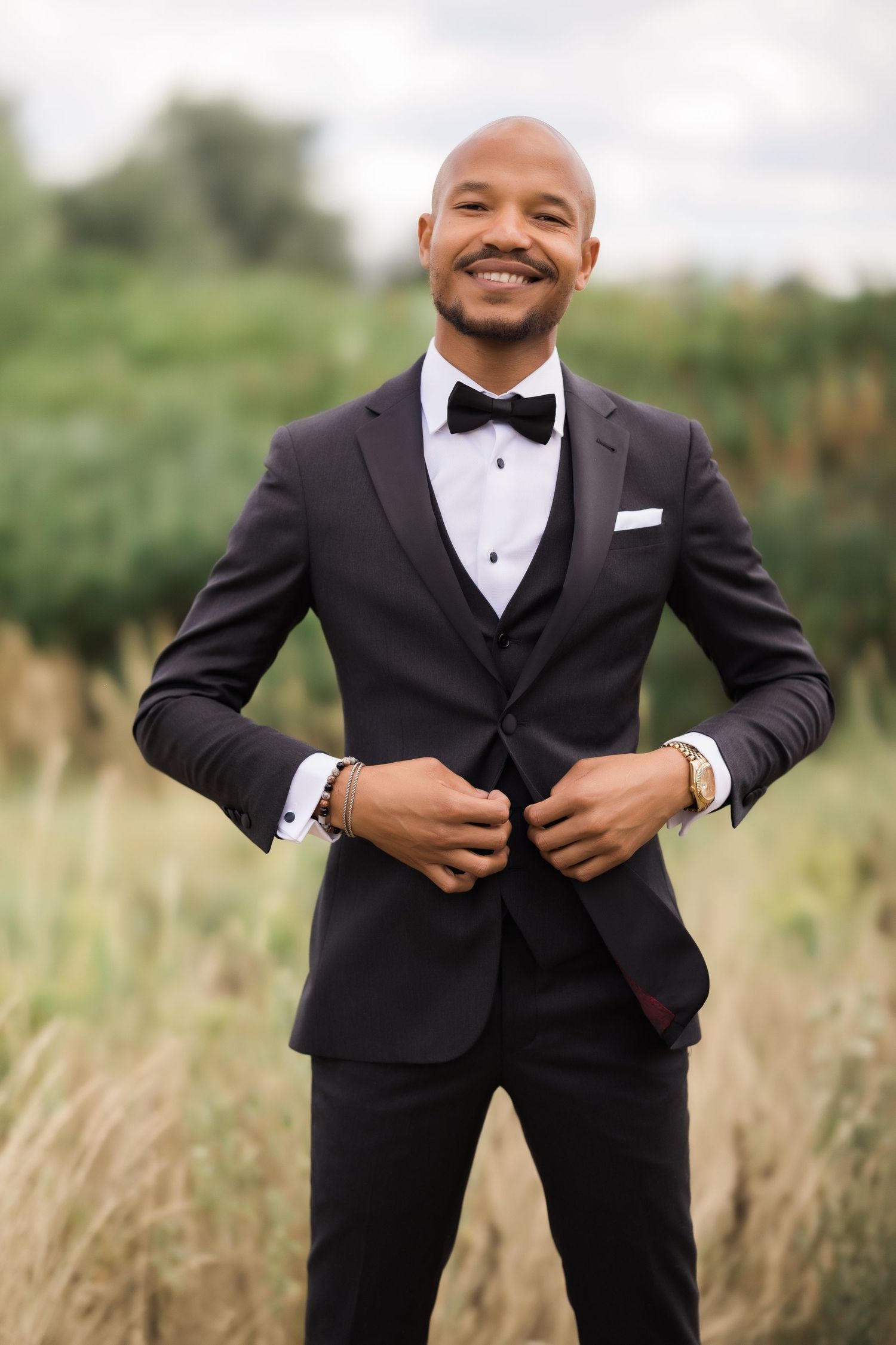 Groom buttons tuxedo jacket while standing in field of tall grass.
