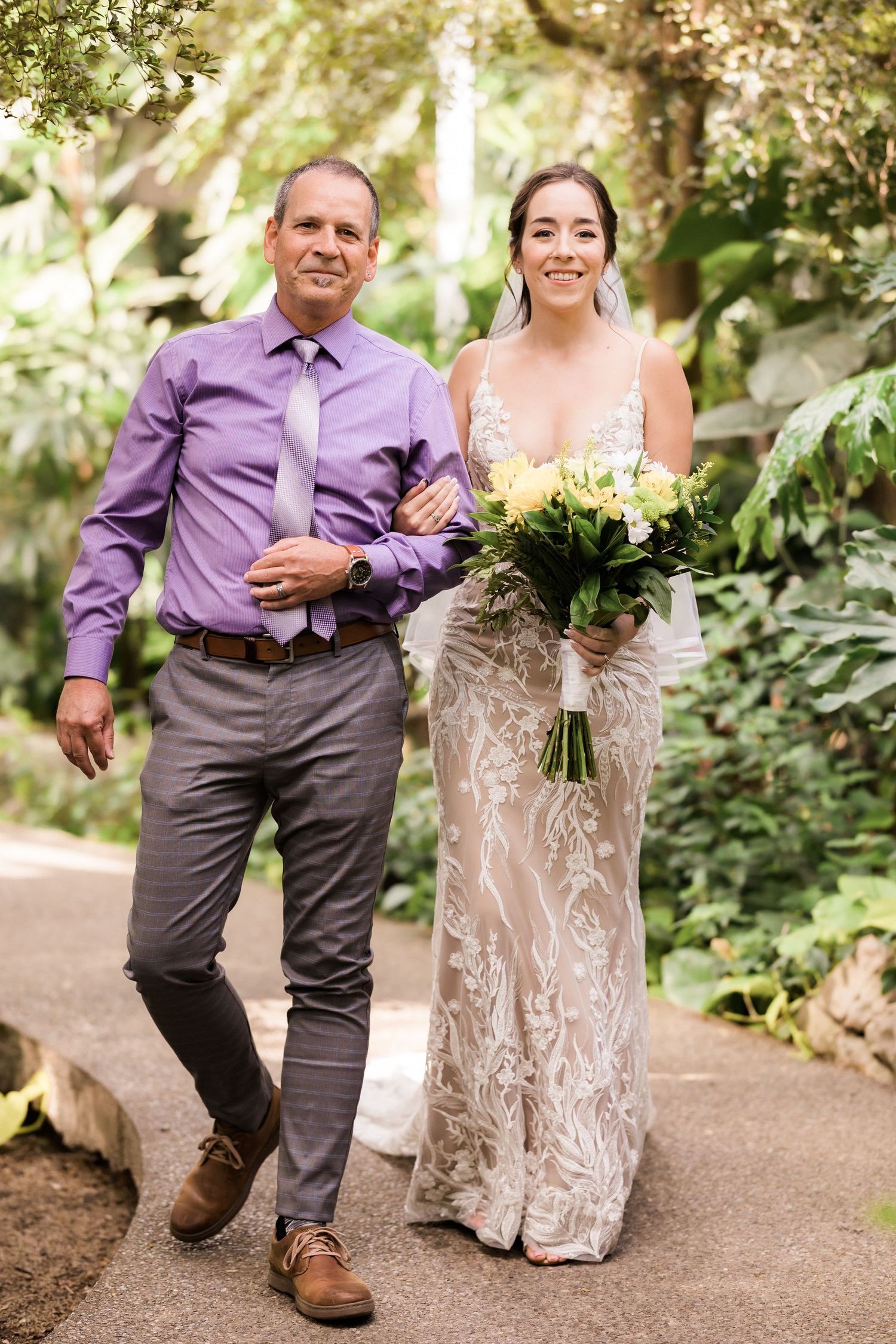 A couple in wedding attire walking along a garden path with lush greenery.