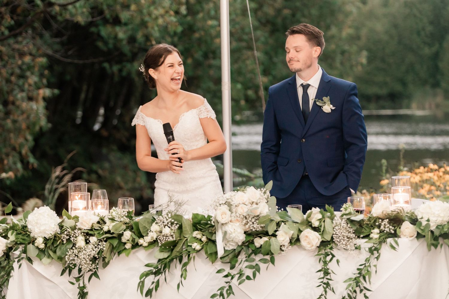 Couple stands behind decorated wedding table with white floral arrangements.