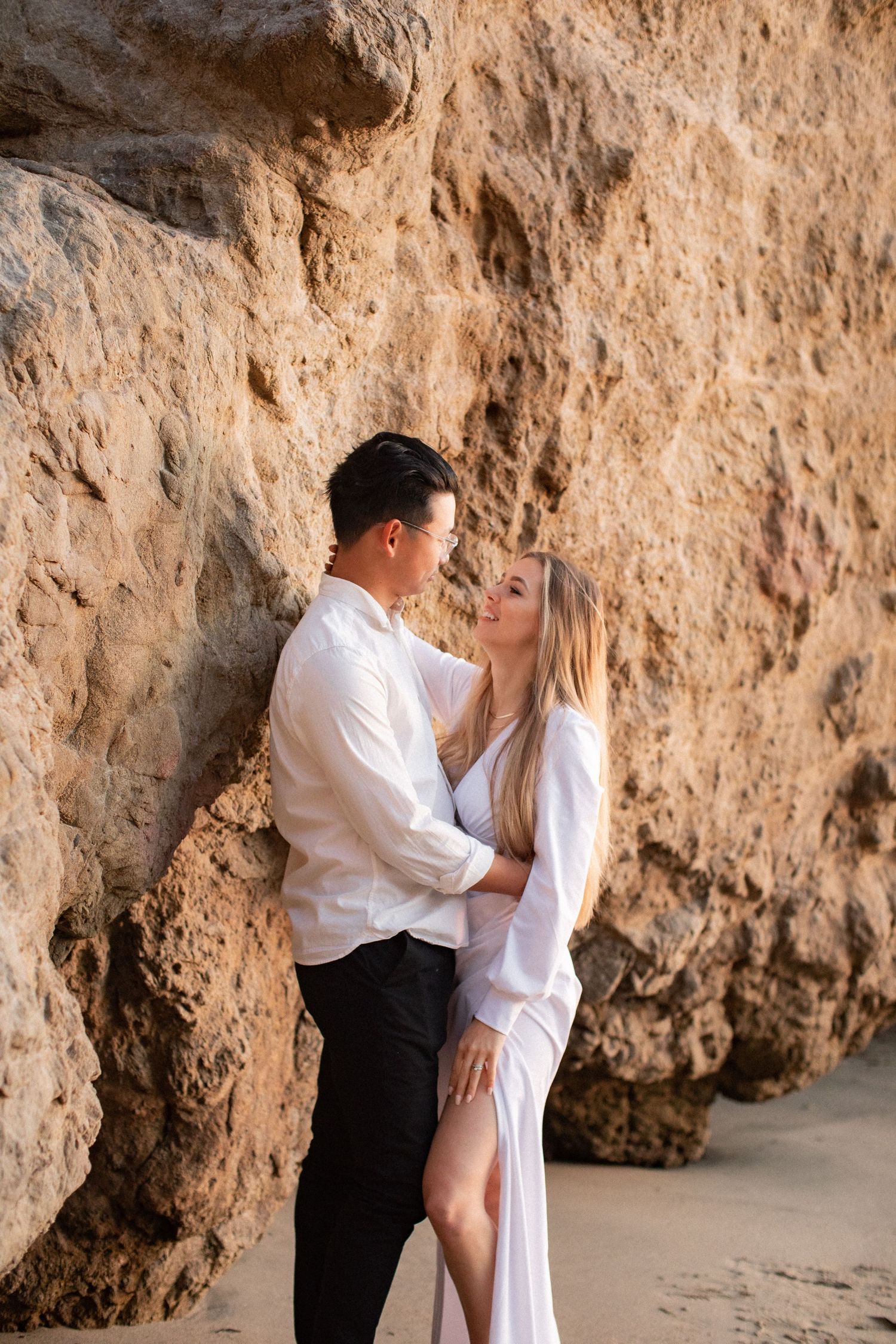 Couple poses intimately against textured limestone cliff face at beach.
