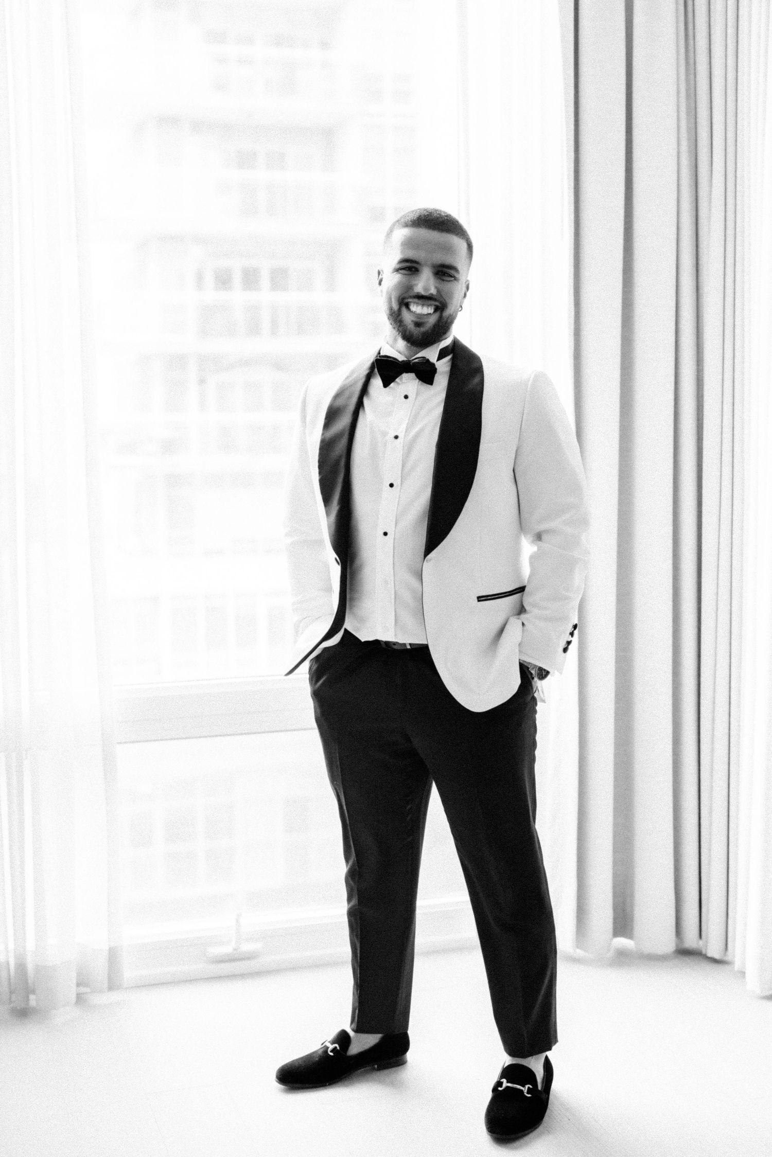 A groom in a white tuxedo jacket and black bow tie smiles in a bright room.