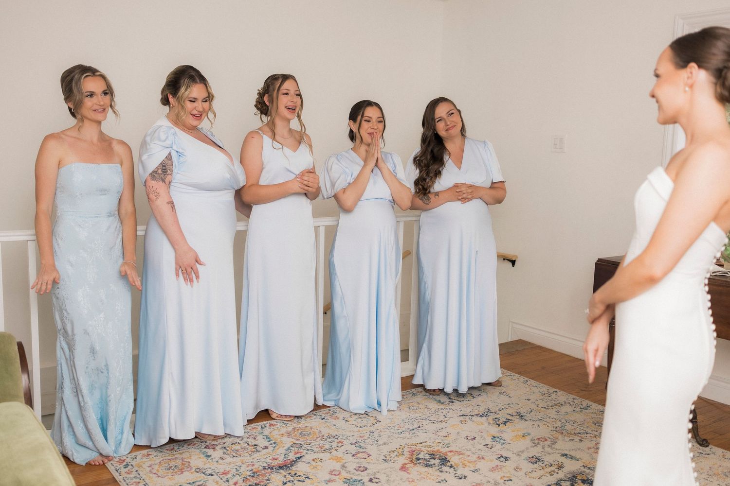 Bridesmaids in pale blue floor-length dresses react with joyful expressions during a wedding preparation moment.