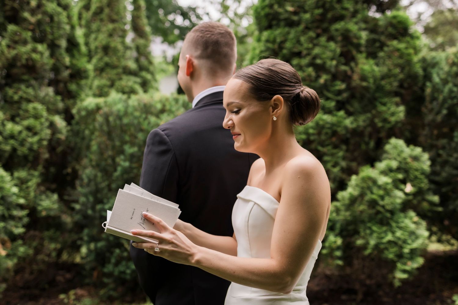 A newlywed pair stands together on a green lawn surrounded by tall evergreen trees.