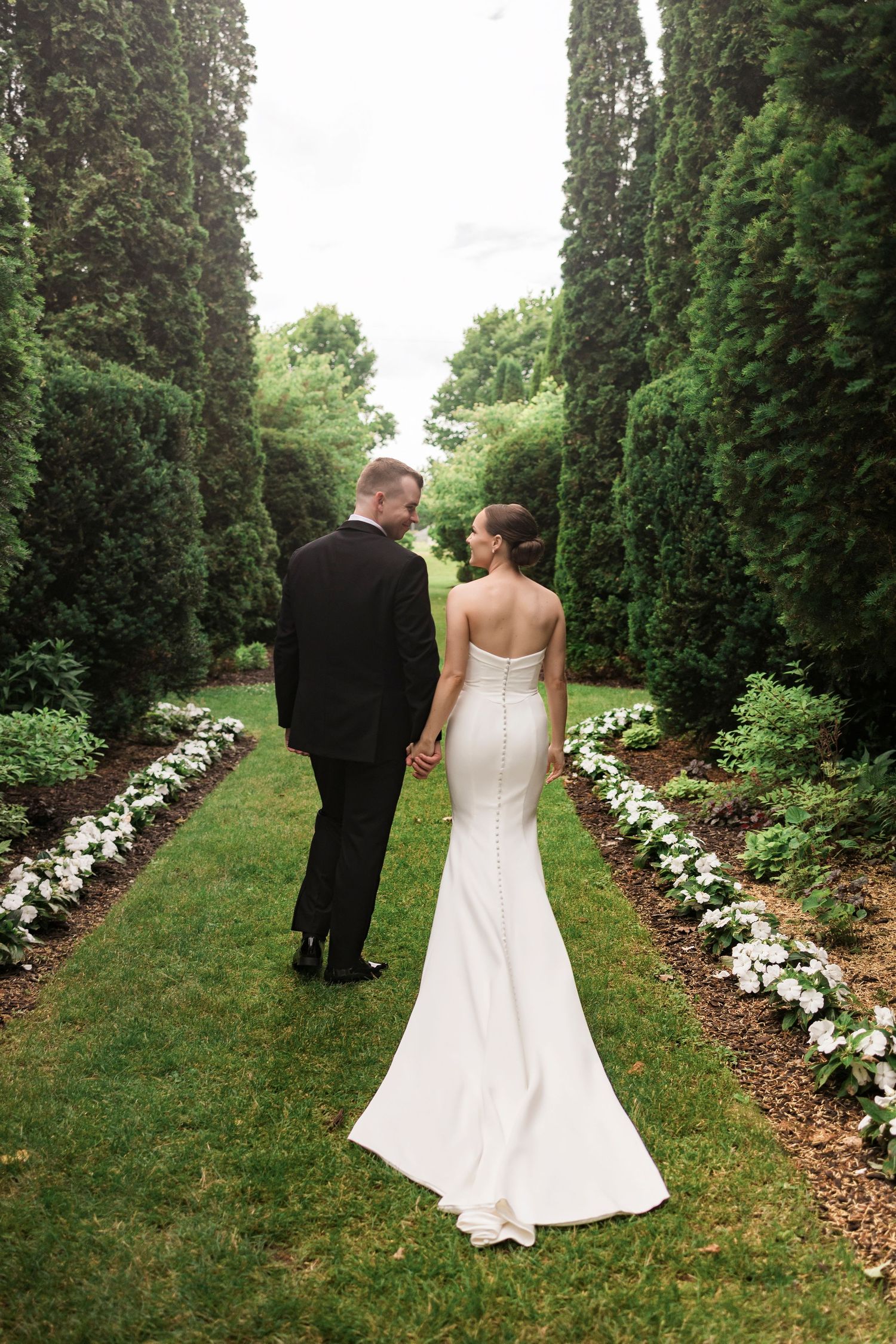 Elegant wedding couple walks down garden path bordered by tall hedges and white flowers.