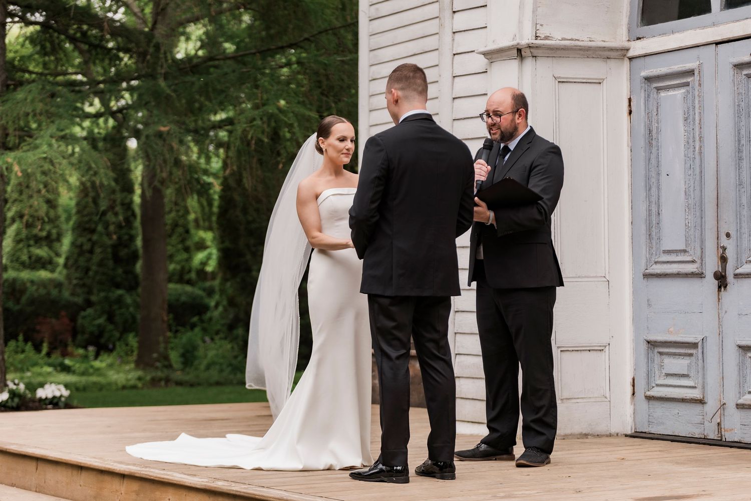 A wedding ceremony takes place on the steps of a white building surrounded by lush greenery.