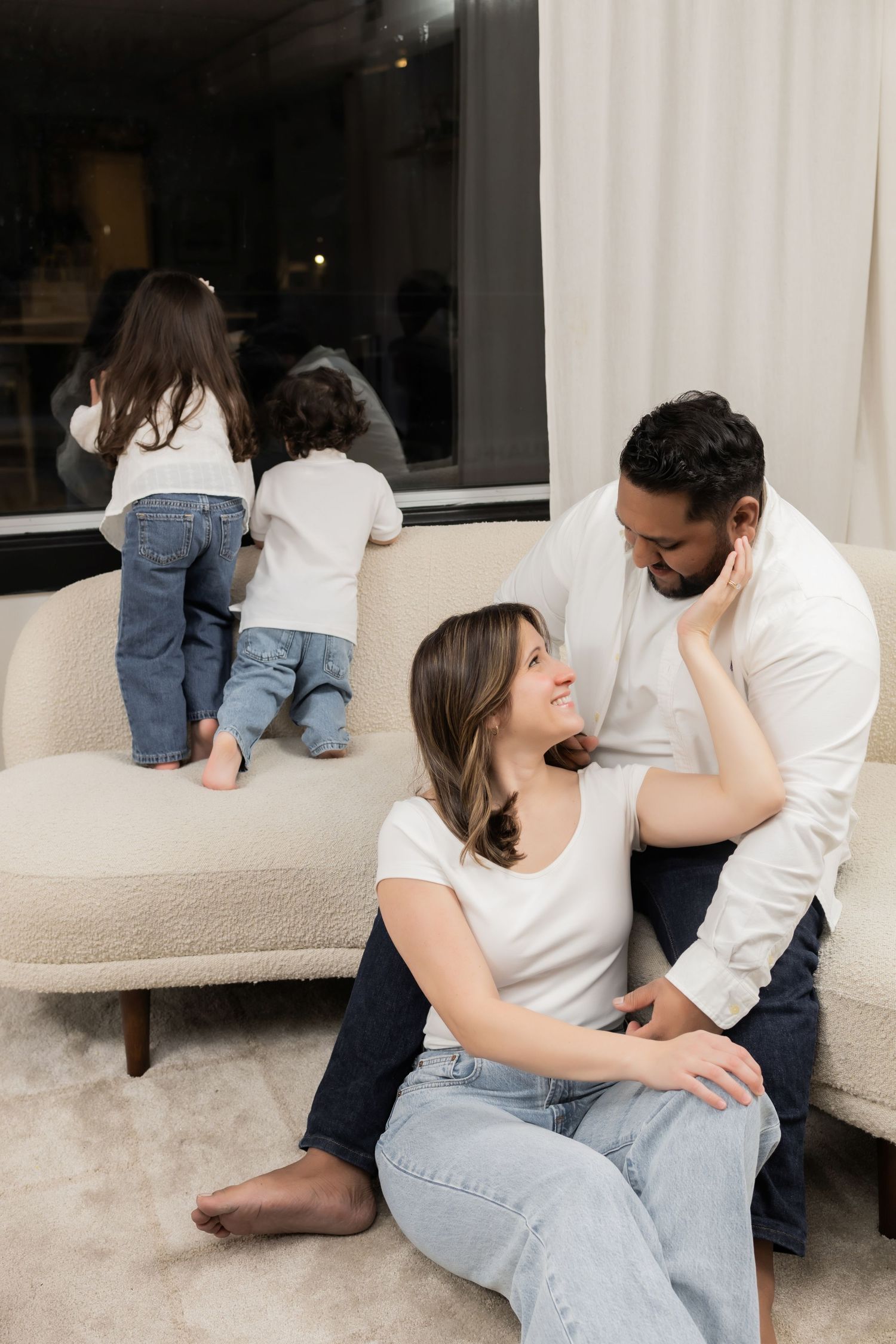 A couple sits intimately together on a couch in a casual living room setting wearing white shirts and jeans.