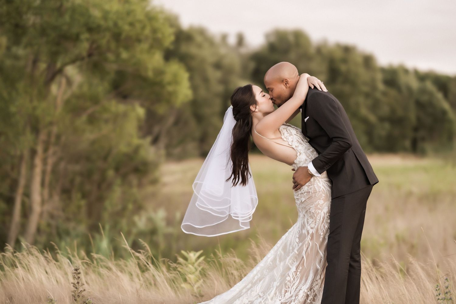 Newlyweds embrace passionately in a rustic field setting with the bride's veil flowing in the wind.