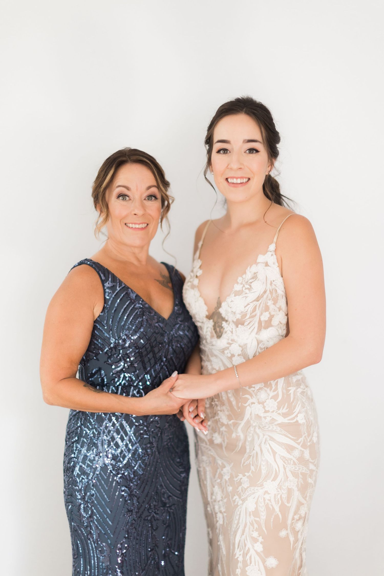 Two women pose together in sequined and lace evening gowns against white backdrop.