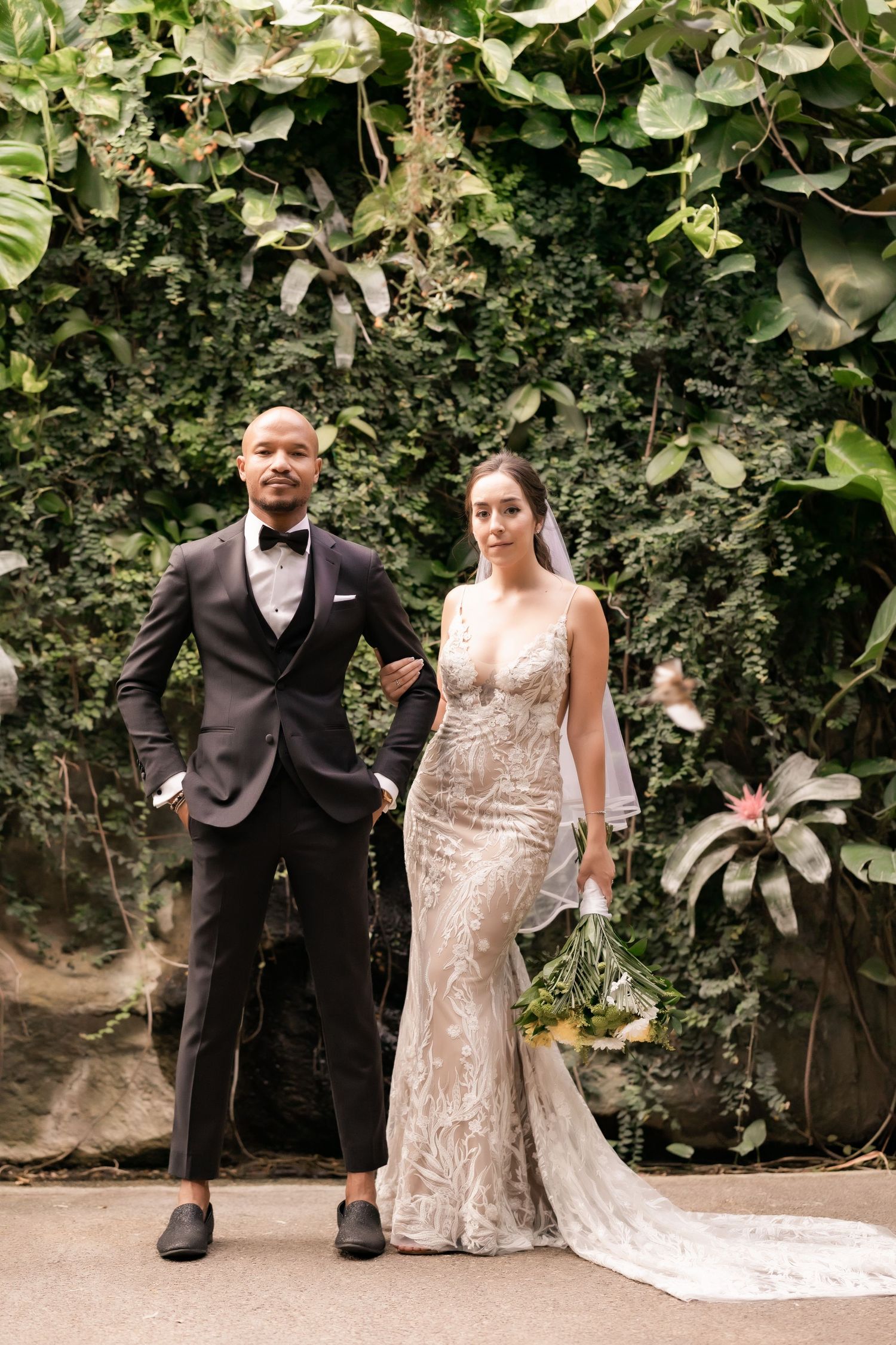 Newlyweds stand together in front of a tropical greenery wall for their wedding portrait.