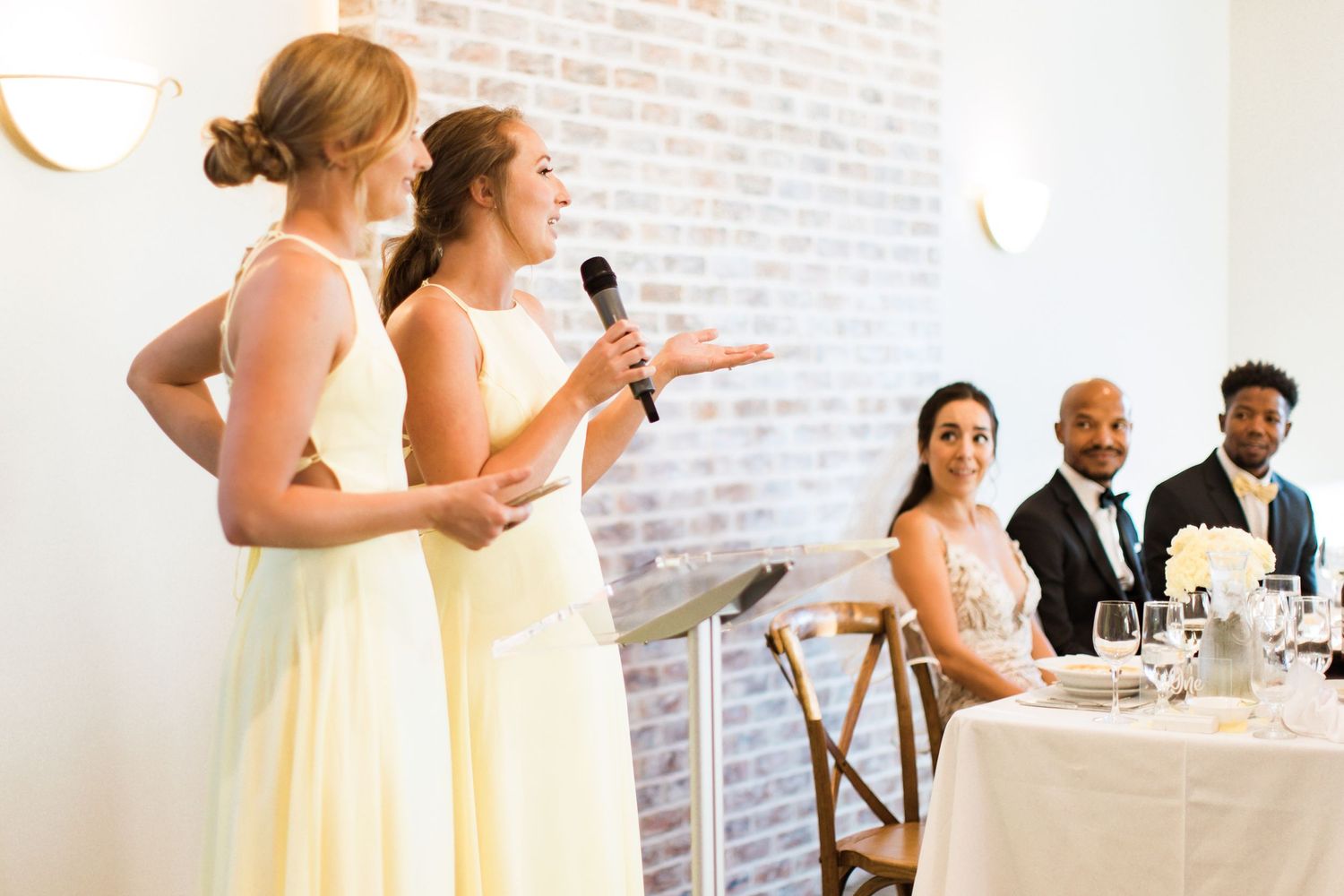 Two bridesmaids in yellow dresses give speeches at a wedding reception with guests seated at tables.