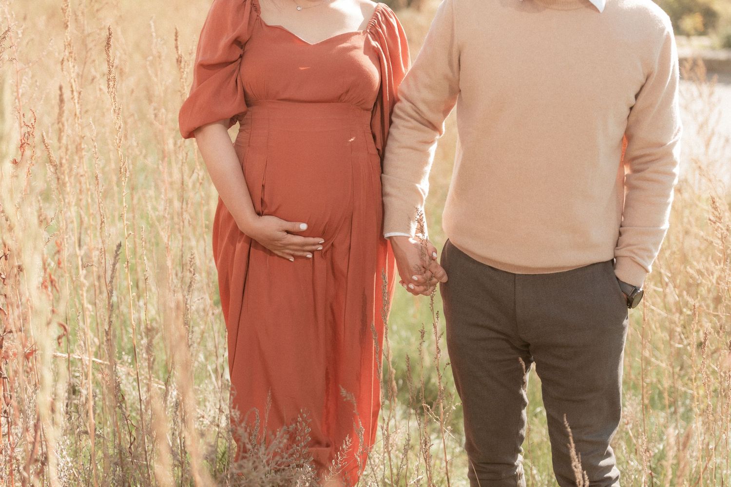 Casual maternity photo of couple holding hands in sunlit meadow with dried grasses.